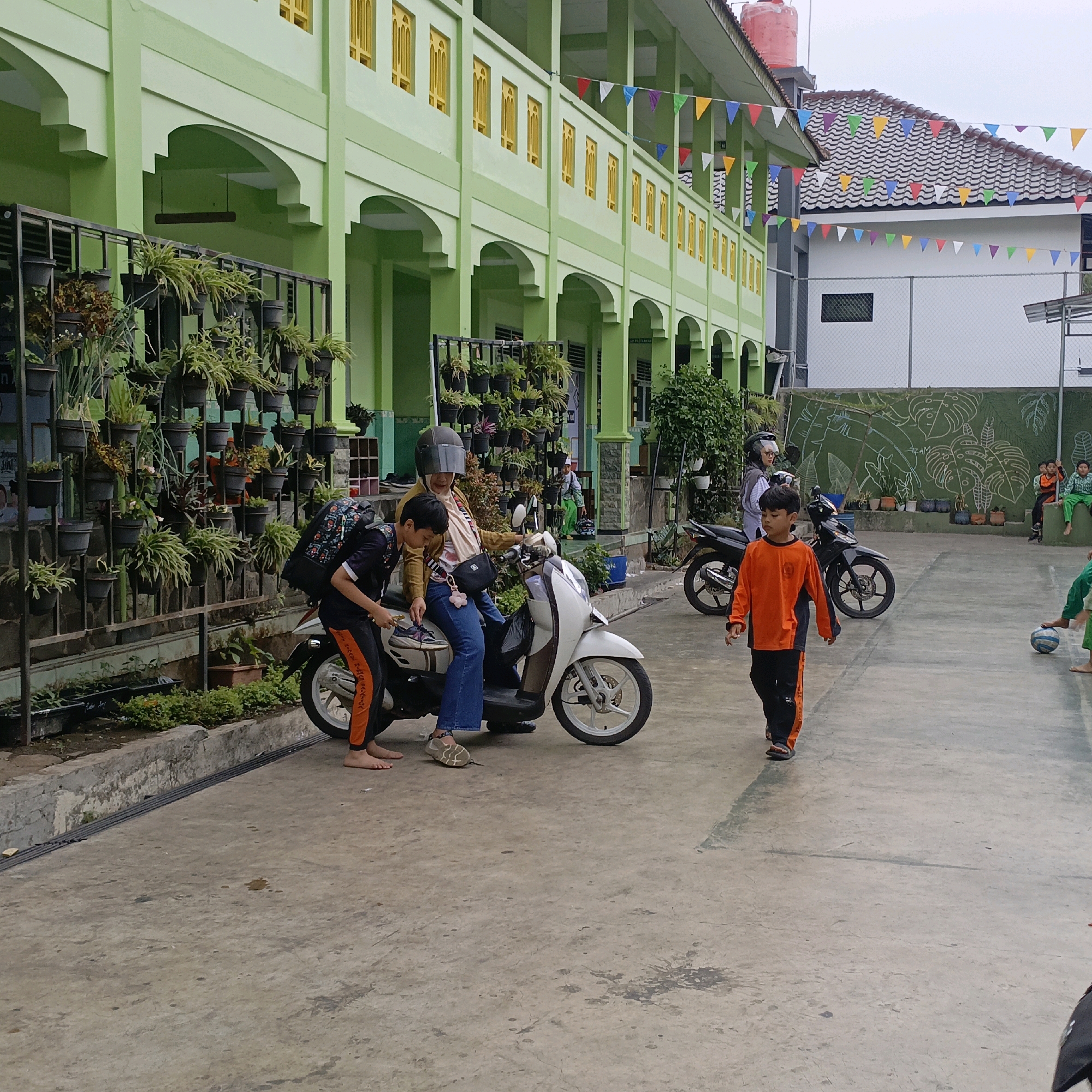 The image depicts a scene outside a light green building with yellow-trimmed windows, likely a school in Salatiga, Indonesia. In the foreground, two young boys are by a white motorcycle. One boy, wearing a backpack, is bending down near the motorcycle's front wheel, and the other boy, in an orange and black outfit, walks away from the motorcycle. A woman wearing a helmet and a light brown jacket is seated on the motorcycle. Behind them, another black motorcycle is parked. The building's exterior features hanging plants in pots on a metal rack. In the background, a chain-link fence surrounds a white wall adorned with a chalk drawing of tropical leaves. Several children are visible near the fence, some seated and others standing, with one group interacting with a soccer ball. Colorful triangular flags are strung across the area, decorating the building's facade and rooftops.