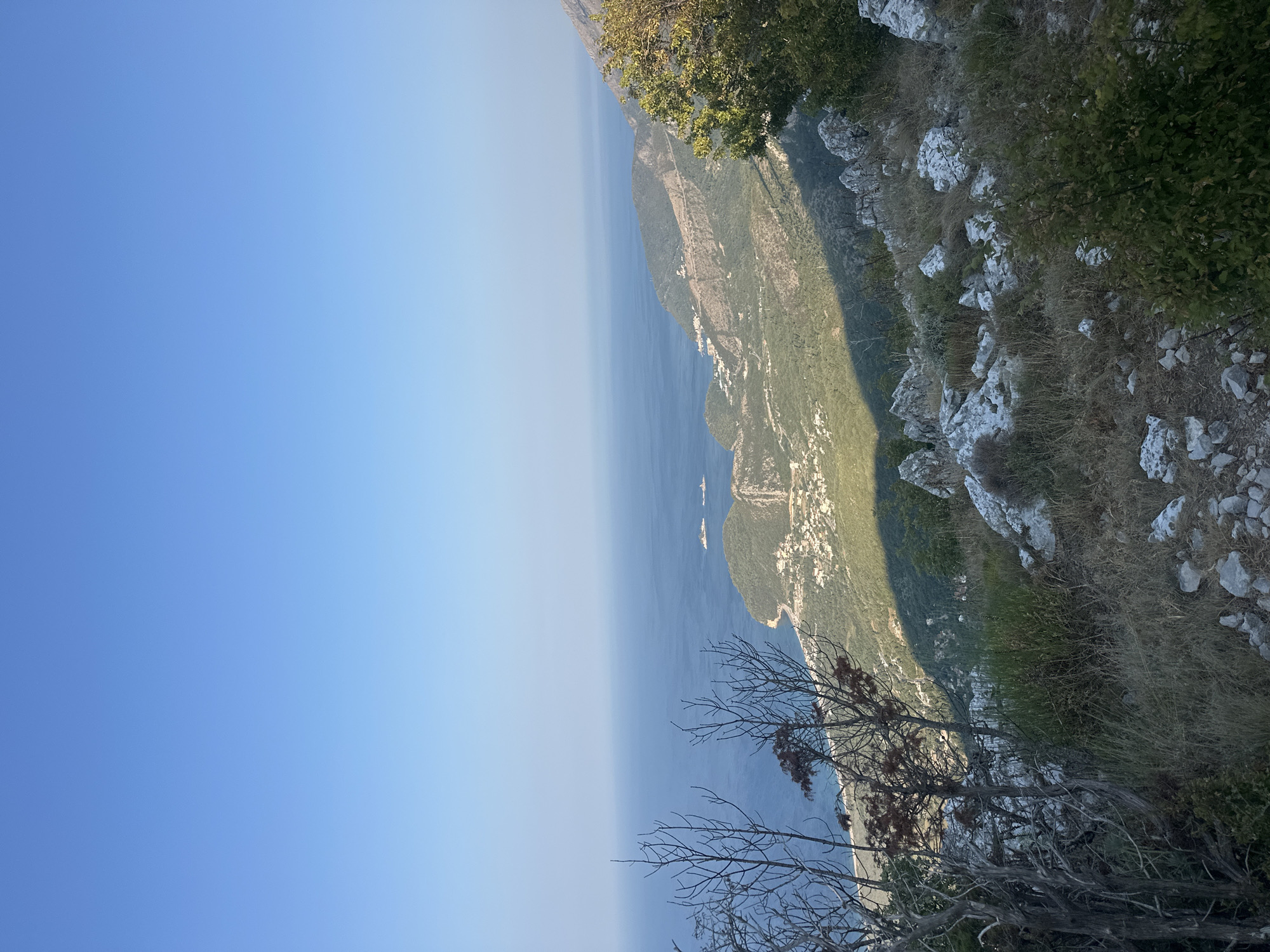 This image captures a panoramic view from a mountainside overlooking the Adriatic Sea near Gluhi Do, Montenegro. In the foreground, the rugged terrain is characterized by dry, sparse vegetation, including bare branches of trees and dry grasses, interspersed with weathered grey rocks. A clear, bright blue sky dominates the upper portion of the image.
The mid-ground reveals a coastal landscape where the deep blue of the sea meets a verdant, hilly shoreline. A small town or village is nestled along the coast, with buildings appearing as white or light-colored structures. The coastline curves, with peninsulas and bays visible. In the distance on the water, two white objects, possibly boats or structures, are discernible.
The sunlight casts a distinct shadow across the hillsides, indicating the time of day, likely late afternoon or early morning, with the sun positioned to the right of the frame. The overall impression is one of a remote, scenic coastal environment, showcasing the natural beauty of the Montenegrin coastline. No people or animals are visible in the image.