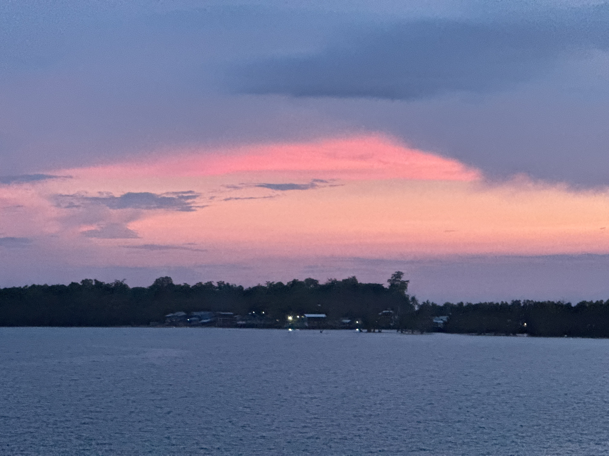 The image depicts a twilight scene over water, likely a bay or inlet, with a landmass visible in the distance. The sky is dominated by hues of pink, orange, and purple, indicative of a sunset or sunrise. There are clouds present in the sky, some darker and more defined, while others are lighter and blended with the colorful sky.
The landmass across the water appears to be an island or a coastal area with dense vegetation. Some buildings or structures are visible along the shoreline, with a few lights suggesting human habitation.
The water is calm with a slight ripple texture and reflects the colors of the sky.
Based on the prompt mentioning Davao City, Philippines, it's plausible that the scene could be near the coast of Davao City, though without specific landmarks, it's impossible to pinpoint the exact location.