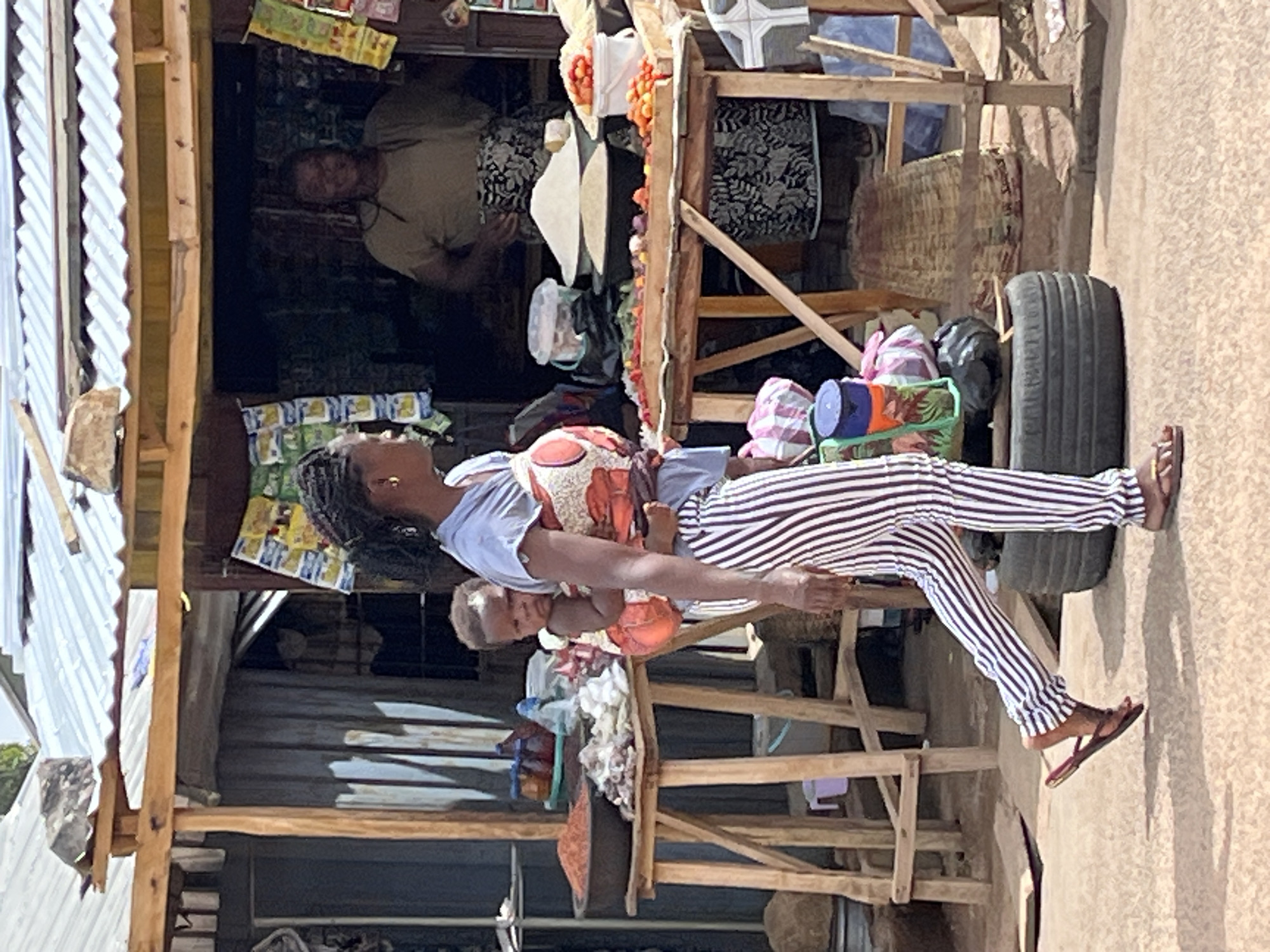 A medium shot shows a woman holding a baby in front of a small wooden stall in Jos, Nigeria. The woman wears white and black striped pants, a light blue shirt, and a patterned orange and white top for carrying the baby. The baby is fair-skinned and light-haired. The stall is made of rough wooden planks and a corrugated metal roof. It displays various items for sale, including tomatoes, containers, and baskets. Another person can be seen sitting inside the stall. A tire leans against the stall on the right side of the frame. The ground is sandy and the lighting is bright.