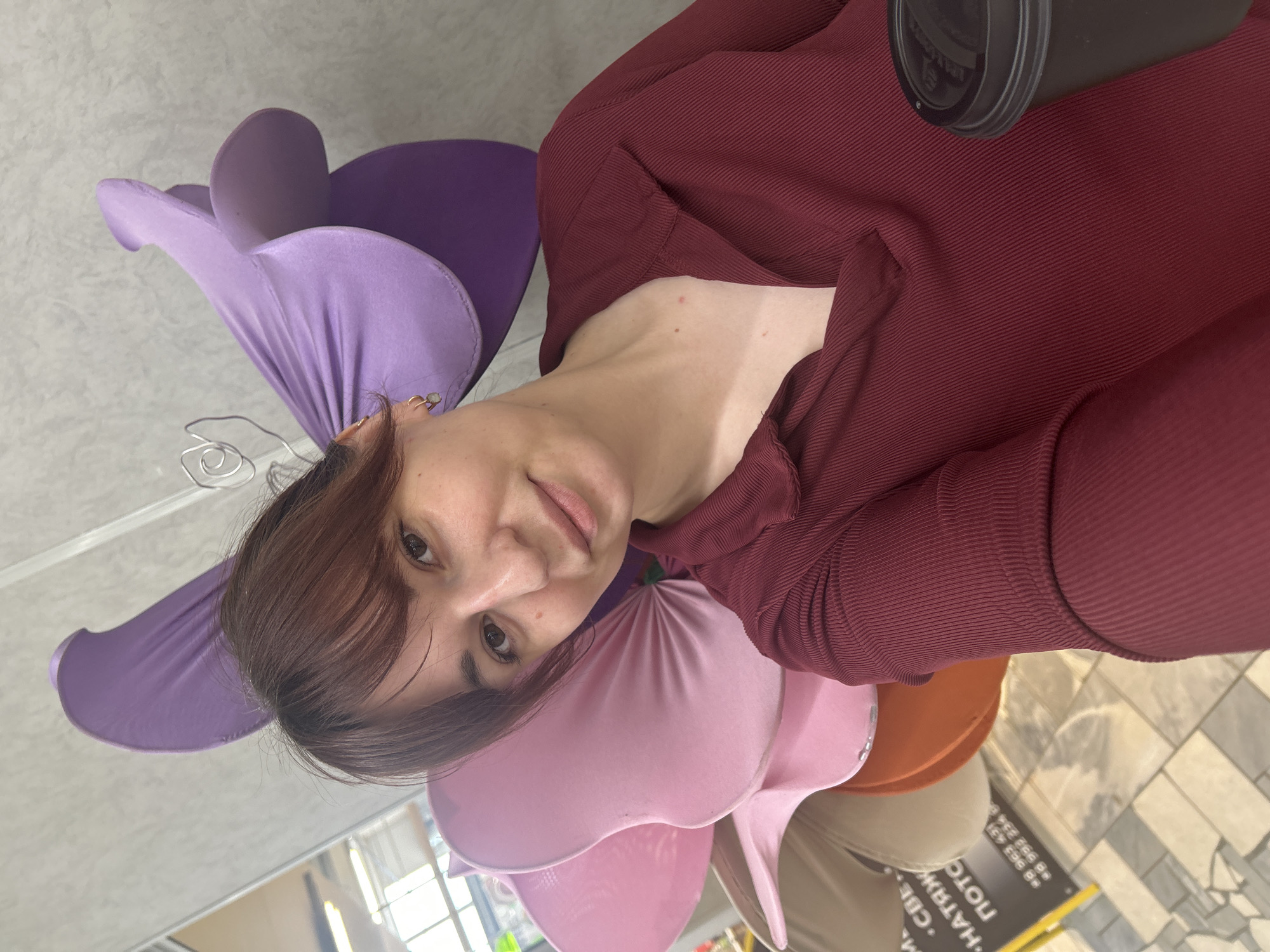 A young woman with brown hair and bangs, wearing a dark red ribbed long-sleeved shirt with a collar, is captured in a close-up selfie. She has small gold hoop earrings in her left ear, smiles faintly, and looks directly at the camera. She holds a black disposable coffee cup with a lid in her right hand, resting against her body. Behind her, large, fabric-covered artificial flowers are arranged, with a purple one positioned behind her head and a pink one behind her left shoulder, and additional tan and orange flowers visible further down. The background reveals a light grey textured wall, a partial reflection of an interior space with ceiling lights, and a dark commercial sign with Cyrillic text. The visible text includes fragments like "СВЕ", "НАТЯЖ", "ПОТО", and phone numbers "+8 963 431" and "+8 992 234". This Cyrillic text, specifically "НАТЯЖНЫЕ ПОТОЛКИ" (Stretch Ceilings), indicates a business related to interior finishing. A yellow stand is visible near the bottom left of the sign, and the floor is composed of light-colored rectangular tiles. The scene is located in an indoor commercial setting within the city of Chaikovsky, Russia.