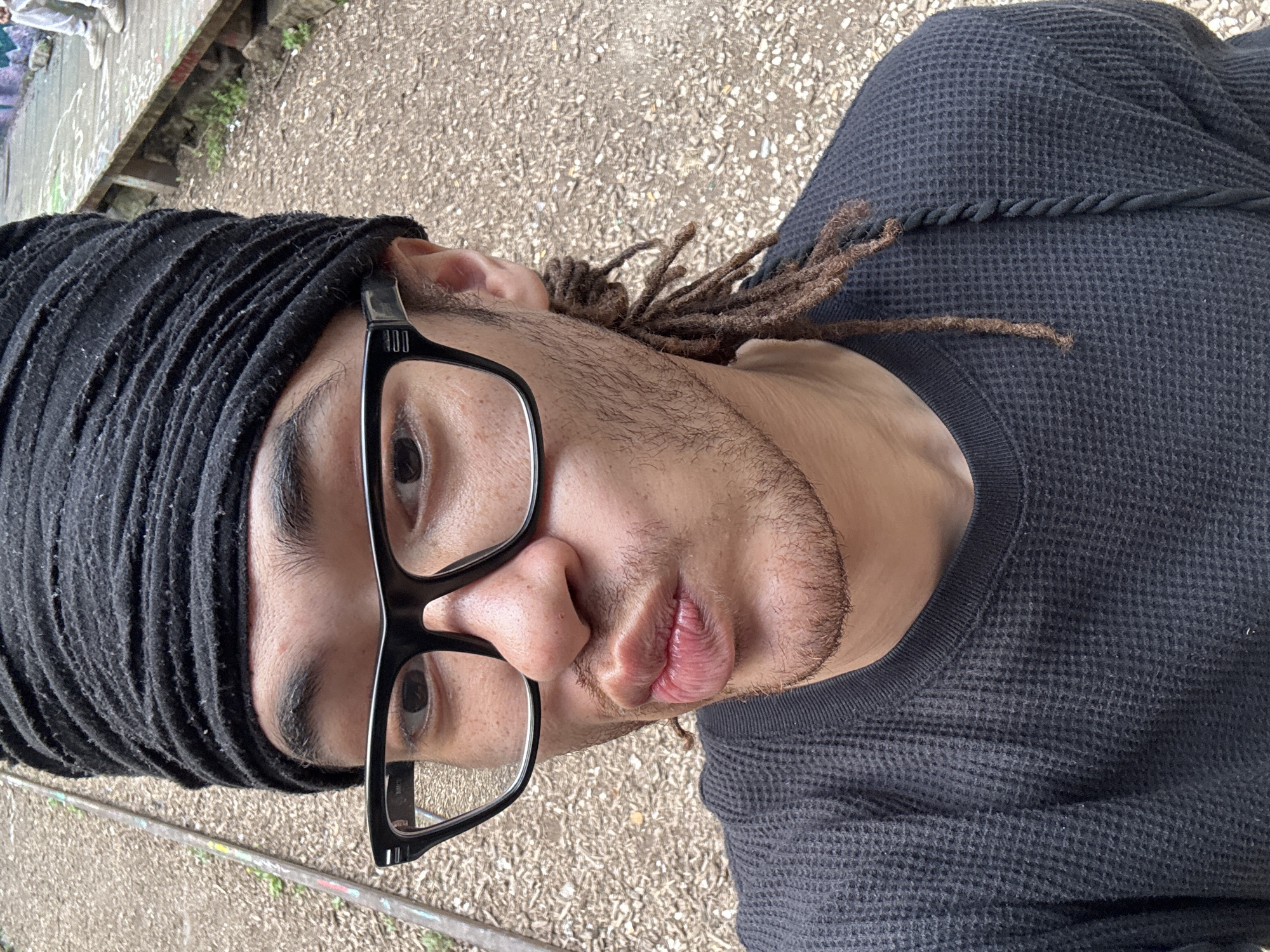A close-up, tilted shot depicts a person with dark skin and dreadlocks, some loose around the face, wearing a black knit cap and black-framed glasses. The person is also wearing a dark, waffle-textured shirt. The background appears to be an outdoor setting with a textured ground surface and a raised edge with vegetation. The location in Paris, France, is not discernible from the given information.