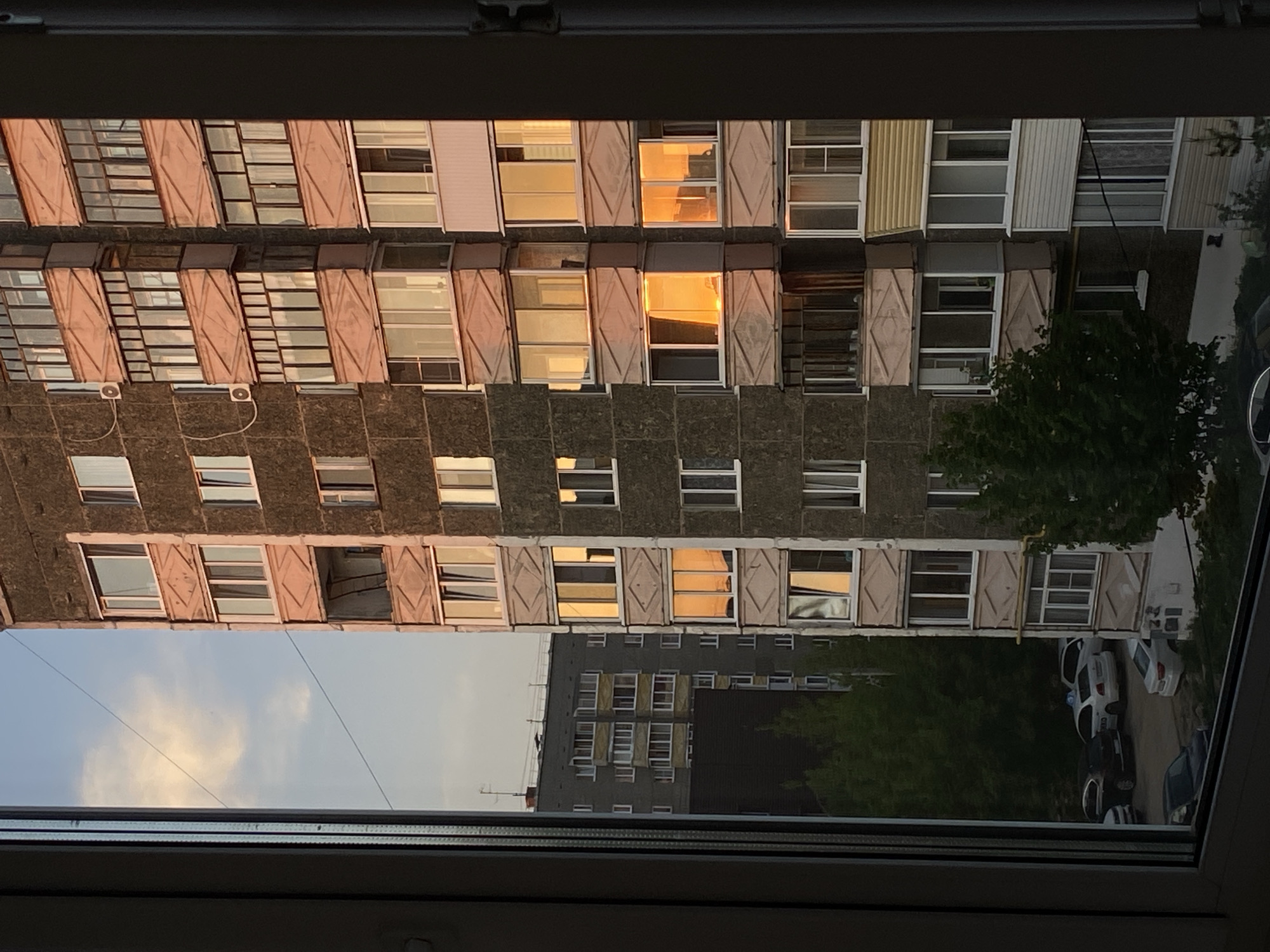 The image depicts a view from a window in what appears to be an apartment building in Votkinsk, Russia. The primary focus is on the exterior view, showing another apartment building across the street. This building is multi-storied with a gray facade and several windows, some reflecting the golden light of either sunrise or sunset. Some of the windows have light-colored shutters or window coverings. A tree is visible on the right side of the frame, partially obscuring a parking area where several cars are parked. The sky is visible in the lower left corner. The window frame in the foreground is white and dark.