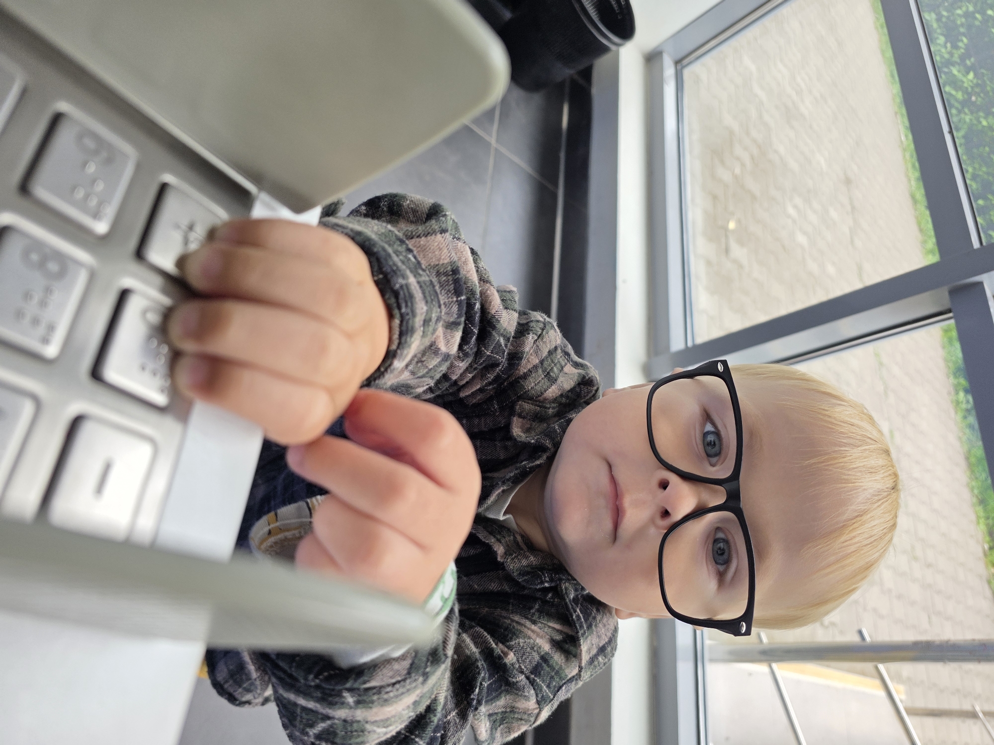 A young Caucasian child with blonde hair and blue eyes, wearing black-rimmed glasses and a green and black plaid shirt, is seated at a keyboard with Braille characters. The child is looking directly at the camera with a neutral expression and has their hands resting on the keyboard. The background shows a tiled floor and a large window or skylight with a view of an outdoor paved area. The scene appears to be indoors, possibly a public building.