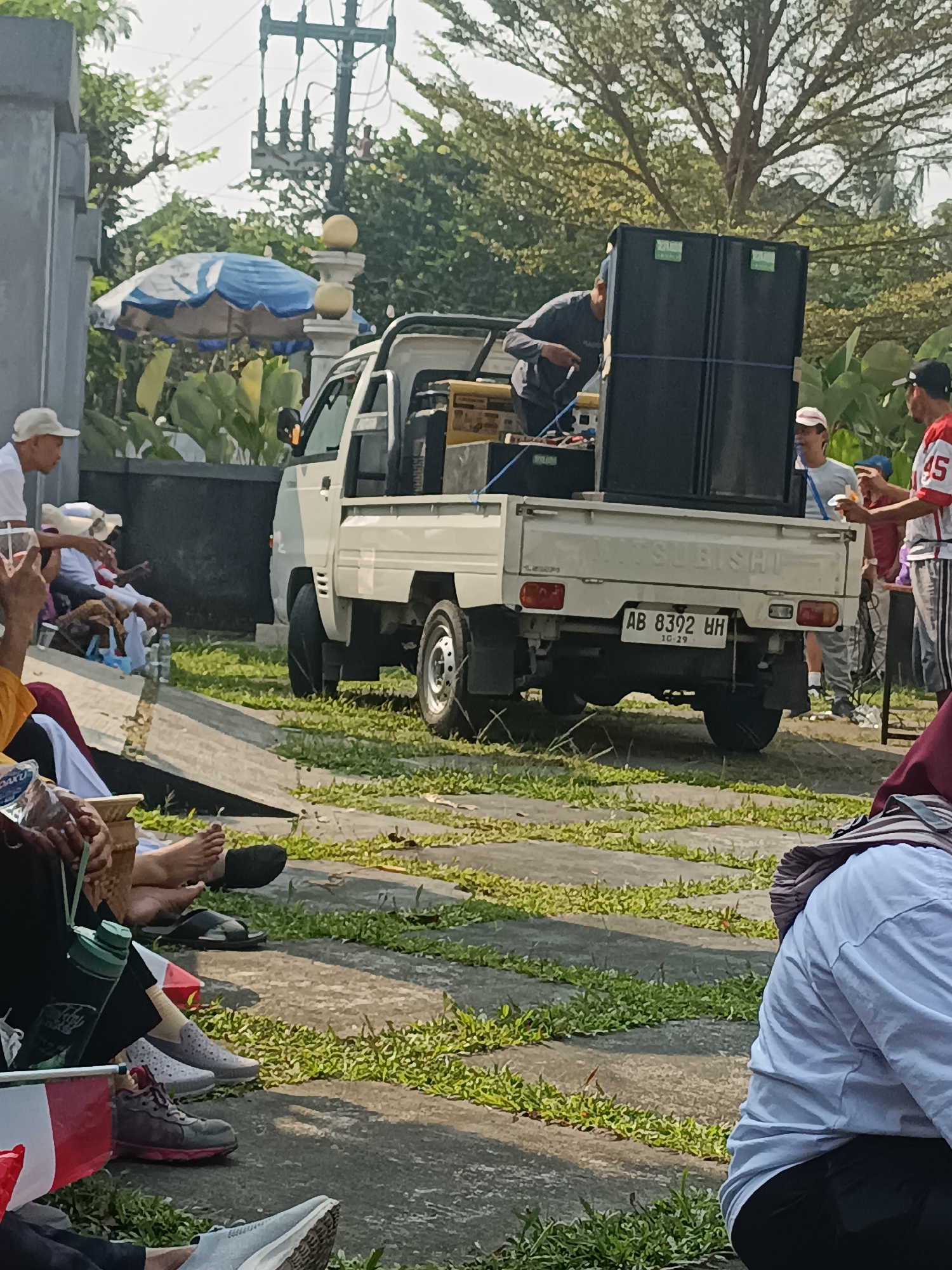 A flatbed truck is parked on a paved area with grass in between the paving stones. The truck is loaded with large speakers and audio equipment. A person in a dark shirt is standing on the back of the truck, appearing to be adjusting the equipment. Several people are seated on the ground in the foreground, some with their legs crossed, facing away from the truck. One person in the foreground is wearing a white shirt and black pants. Another person in the middle ground is wearing a red and white shirt. The environment appears to be outdoors, with trees and foliage visible in the background. The truck has a license plate that reads "AB 8392 BH". The scene suggests an outdoor event or gathering where music is being played. This scene is likely located in Sleman, Indonesia, as indicated by the license plate format.