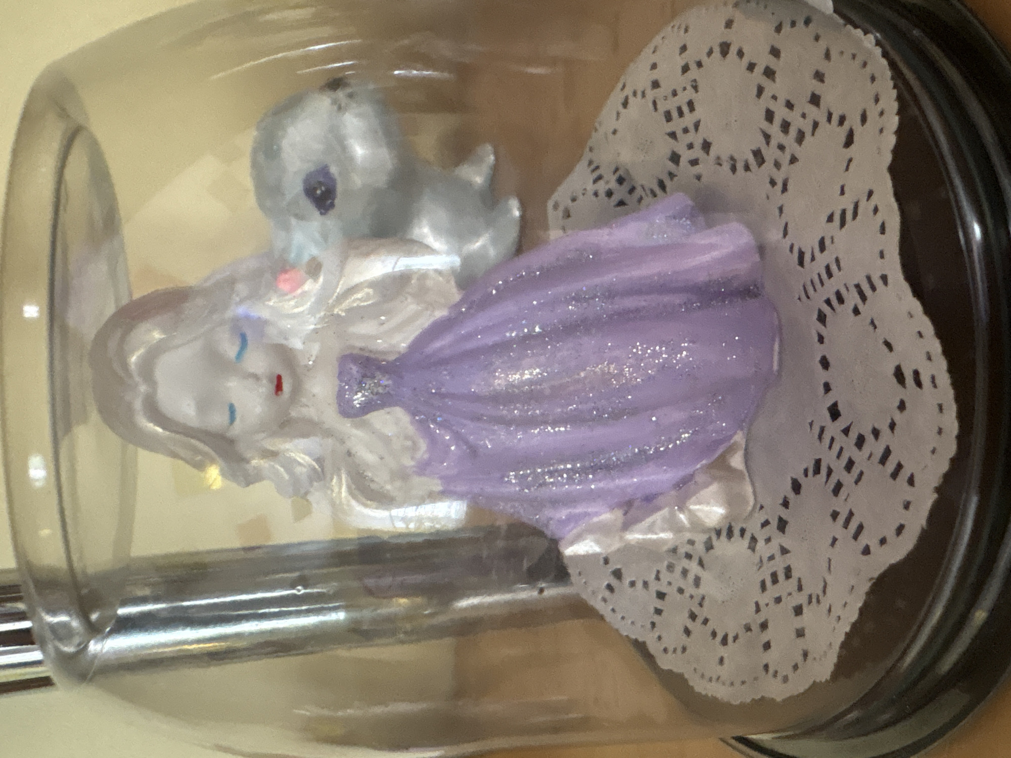 A decorative glass dome with a black base sits on a wooden surface. Inside, a small figurine of a blonde-haired angel with a purple dress adorned with glitter stands on a white paper doily. To the right of the angel is a small, silver-colored figurine of a rabbit. The background is out of focus and appears to be a light yellow color, possibly a wall or curtain. There are no visible people or specific actions taking place within the scene. The overall impression is of a small, whimsical display, likely intended for decoration. The location in Ветрынскі сельскі Савет, Belarus, is not visually determinable from the image.