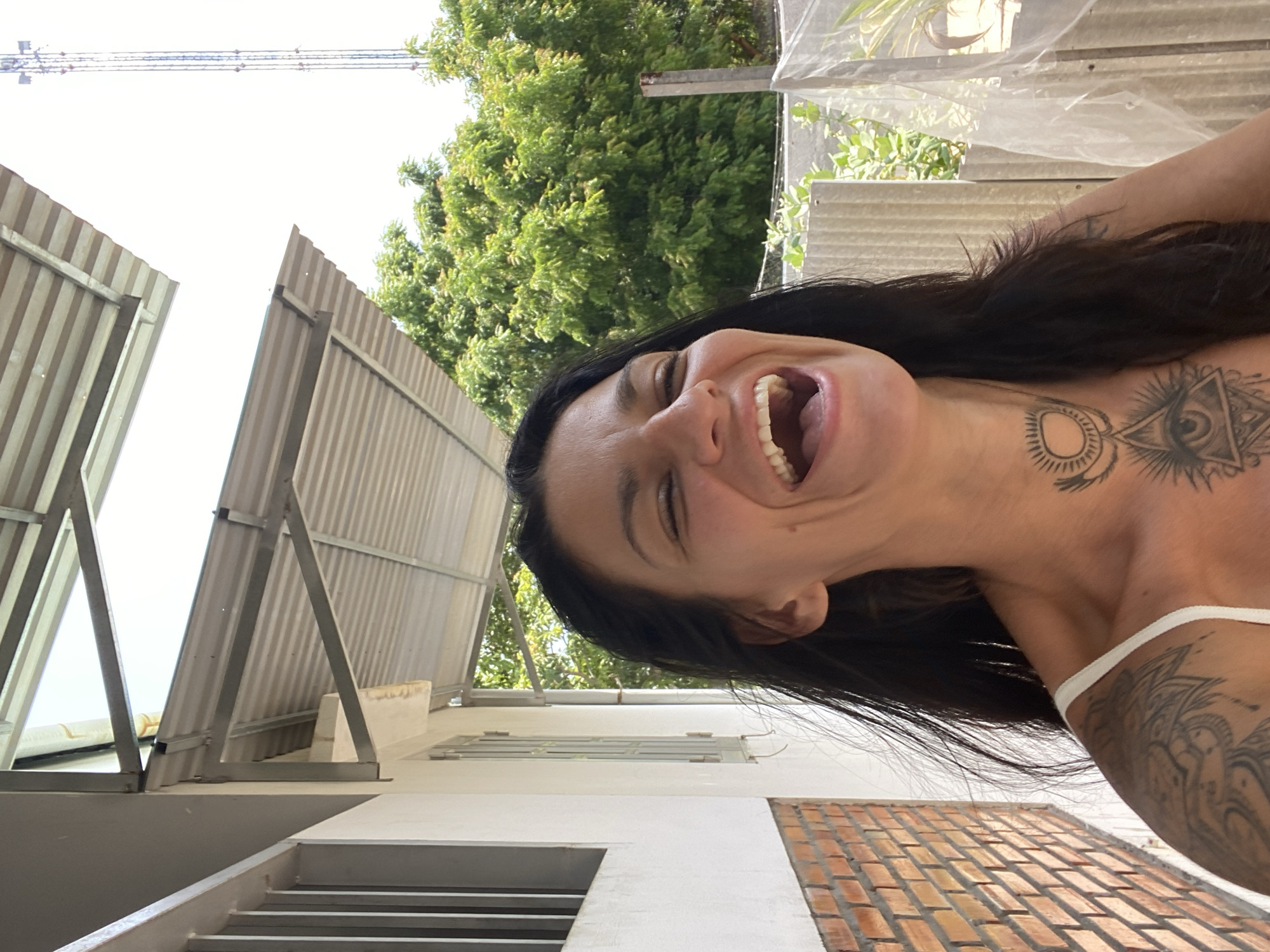 A dark-haired woman with tattoos on her chest, neck, and shoulder is laughing with her eyes closed. She is wearing a white bra strap. The background includes a white building with a brick base, a metal roof structure, lush green trees, and a tall tower in the distance. The scene appears to be outdoors, possibly in a residential or semi-urban area in Mũi Né, Vietnam.