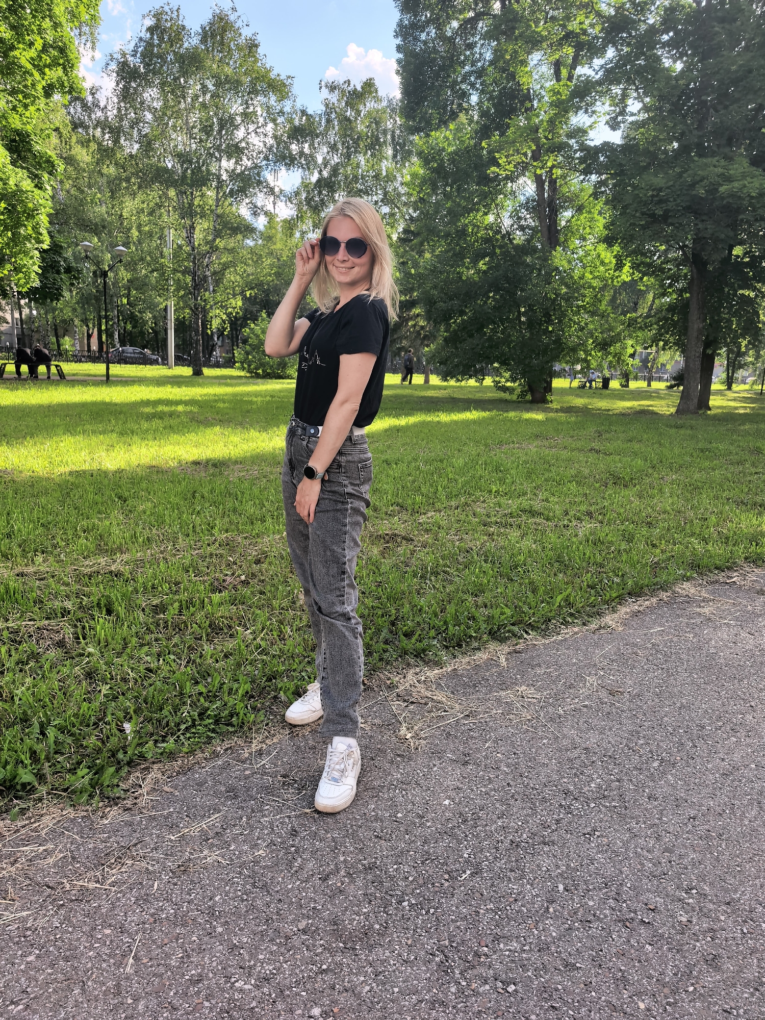 A young, blonde woman with sunglasses is standing on a paved path in a park in Sterlitamak, Russia. She is wearing a black t-shirt, gray jeans, and white sneakers. The woman is looking at the camera and smiling. Behind her, there is a grassy area with trees. The weather appears to be sunny.
