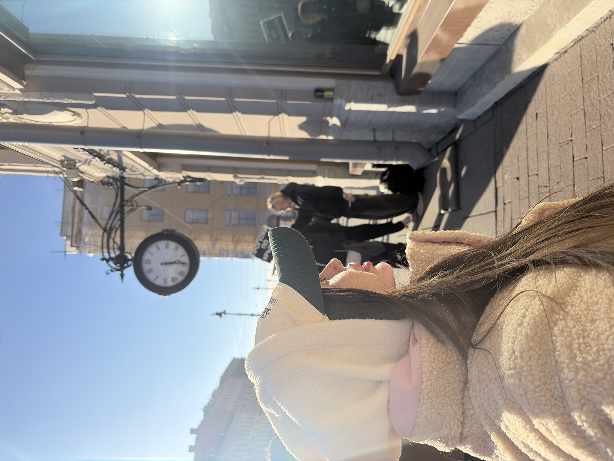 The image shows a woman wearing a white sherpa jacket and a green baseball cap standing on a sidewalk next to a building. In the background, a clock on a lamp post reads approximately 11:55. Behind the woman are two figures walking, one of whom is wearing a mask. The location appears to be Saint Petersburg, Russia, based on architectural styles and street features. The weather seems to be sunny.