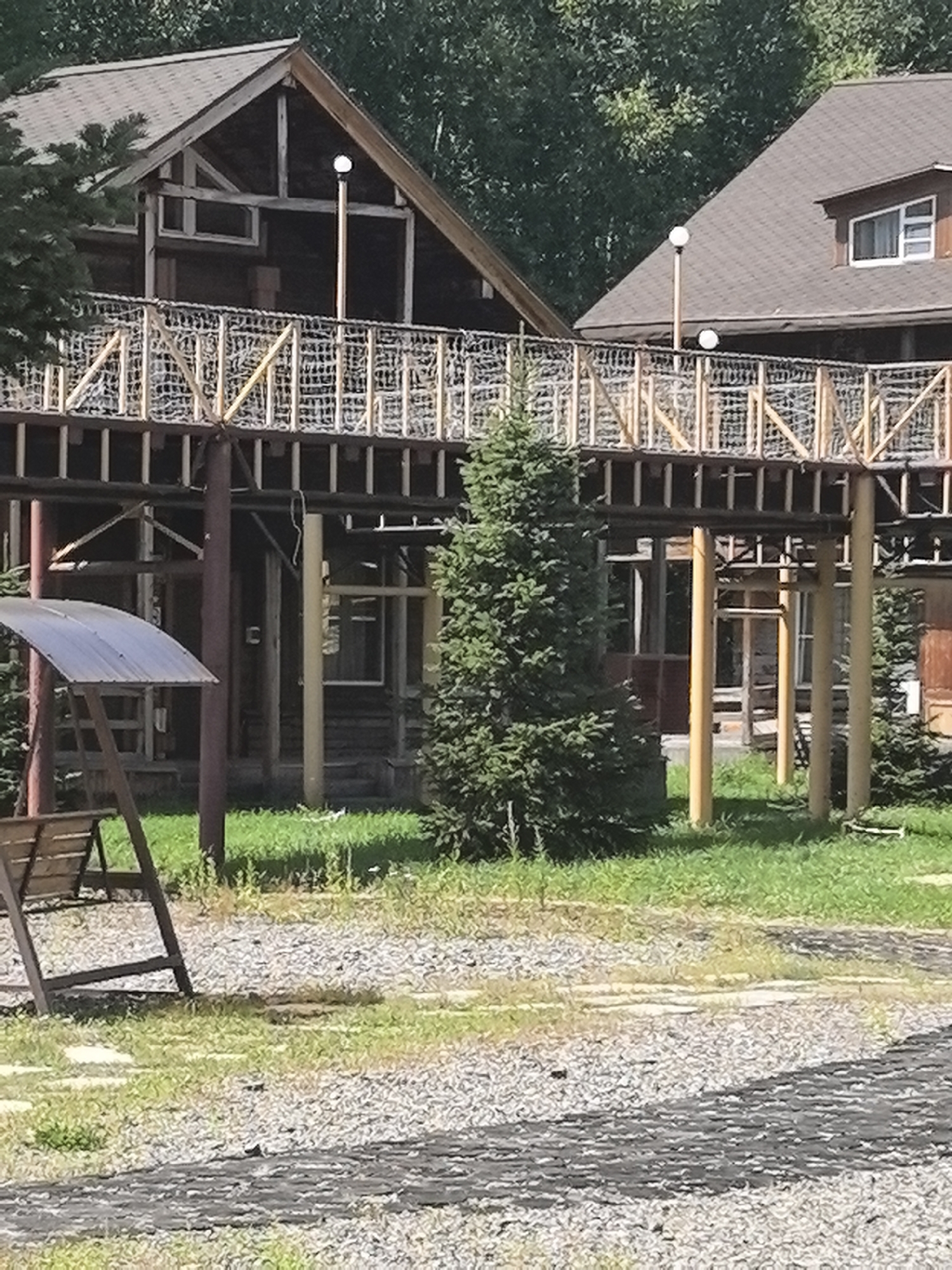The image depicts an outdoor scene in Solnechnyi, Russia, featuring a multi-level wooden structure with a walkway connecting two buildings. The walkway is supported by numerous vertical posts and features a mesh railing. A tall evergreen tree stands prominently in the foreground, positioned to the right of the walkway's support structure. To the left, a wooden bench with a metal canopy is visible, situated on a grassy area. The ground is covered with gravel and patches of grass. The buildings themselves are wooden, with visible rooflines and windows. The overall environment suggests a park or recreational area during daylight.