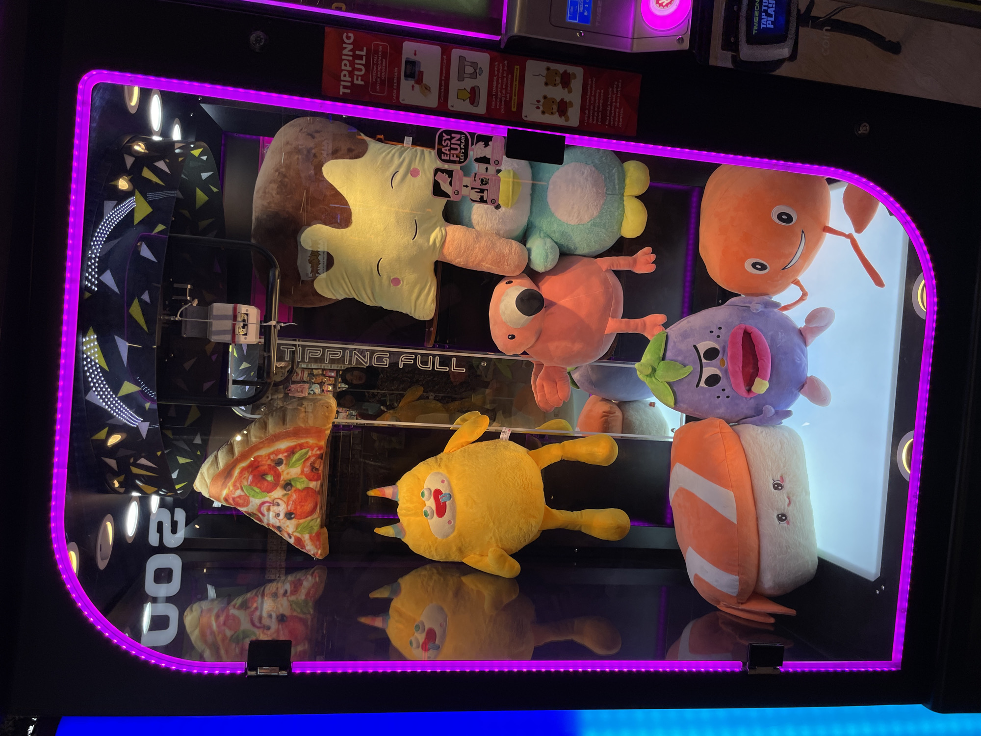 The image shows the inside of a claw machine filled with plush toys. The machine is brightly lit with purple LED lights around the perimeter. In the foreground, from left to right, are plush toys shaped like sushi, an eggplant with a surprised expression, and an orange round character with arms and legs. In the middle row, there are plush toys of a flamingo and a blue and white penguin. At the top, a large brown plush toy with a yellow piece resembling ice cream or a popsicle is visible. A plush pizza slice is also displayed near the top. On the right side of the machine, instructional signs are attached, one of which indicates "TIPPING FULL". The overall environment appears to be an entertainment venue, likely an arcade or shopping mall, given the nature of the claw machine and the surrounding signage. The reflection in the glass shows a blurred background with other arcade machines and possibly people. The text "200" is visible on the top left of the interior of the machine. The machine itself is black with a clear glass front. Based on the presence of the "TIPPING FULL" sign and the overall context, this claw machine is located within the city of Manado, Indonesia, likely in a public entertainment area.