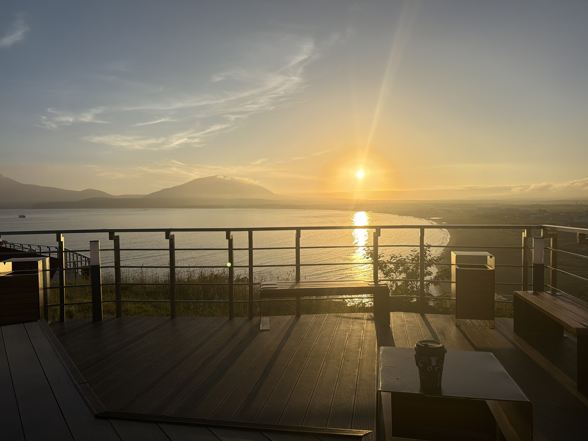 The image captures a serene sunset scene from a wooden deck overlooking a body of water and a distant mountain range. The sun, a bright orb, is setting on the horizon, casting a warm golden glow across the sky and reflecting on the water. Wisps of clouds are scattered across the pale blue sky. In the foreground, a dark wooden deck features several benches, a trash receptacle, and a table with a coffee cup on it. A metal railing separates the deck from a grassy area with tall reeds, beyond which lies the tranquil water. The landscape suggests a coastal location with mountains in the background. The scene appears to be in Yuzhno-Kurilsk, Russia, given the context provided, likely an outdoor seating area at a scenic overlook. There are no people visible in the image.