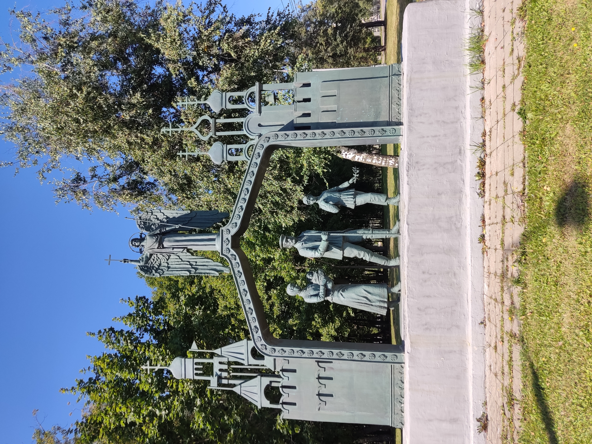 The image shows a bronze monument in a park-like setting with trees and a blue sky. The monument features an arched gateway supported by architectural elements resembling church towers or fortifications. Atop the arch stands a winged angel holding a cross. Beneath the arch, three figures are depicted in a procession: a woman holding a baby, a man with a walking stick, and a young person holding a sprig of greenery. The monument is situated on a concrete pedestal, with a paved walkway and grass in the foreground. The style of the monument suggests a historical or religious theme. This is the monument "The Golden Gate of Vladimir" on Borovitskaya Square in Moscow, Russia.
