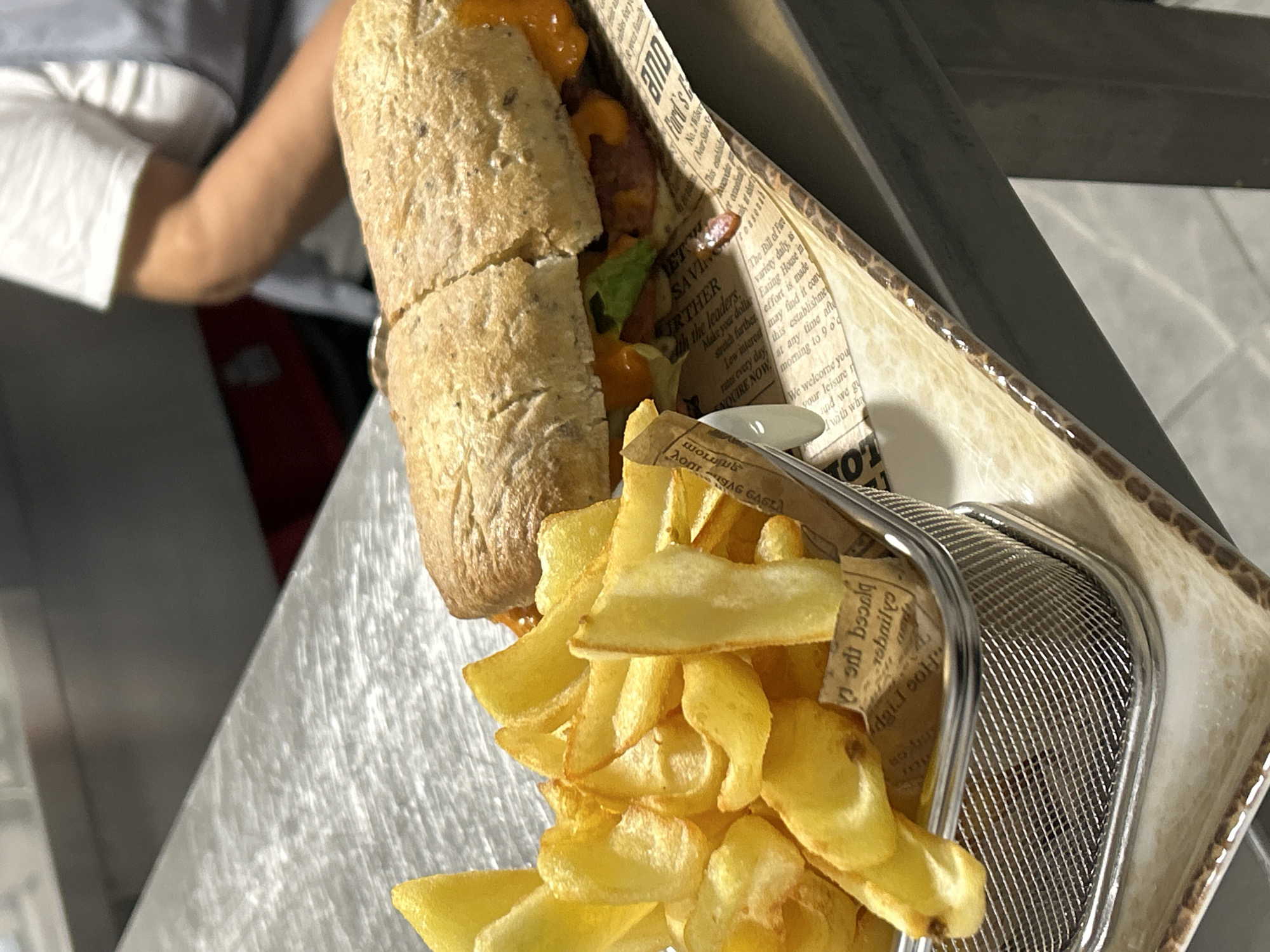 A sandwich and fries are presented in an elevated, angled view. The sandwich, cut into two pieces, features a grainy bread roll filled with what appears to be lettuce, sauce, and a strip of meat. It rests on decorative paper with printed text and patterns. A small, white, oval-shaped container is partially visible, sitting near the sandwich. Next to the sandwich, a metal wire basket is filled with golden-yellow fries. The fries are also partially spilling out of the basket. The food is placed on a metallic surface with a brushed finish. In the background, a blurred figure wearing a light-colored shirt is visible. Location details, such as Burgas, Bulgaria, are unknown.