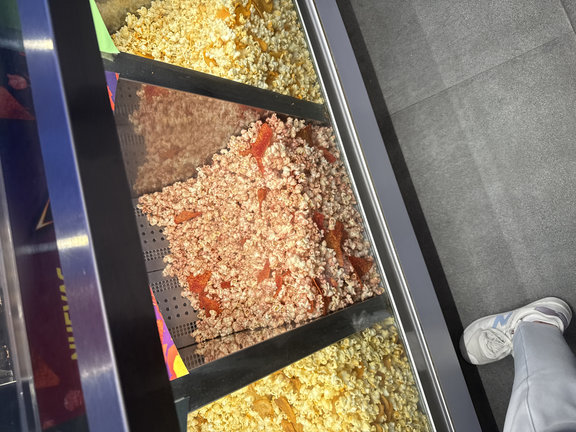 The image shows a display case with three levels of popcorn. The top and bottom levels contain yellow popcorn, while the middle level contains red-colored popcorn with what appear to be tortilla chips mixed in. A person wearing white sneakers and light-colored pants is standing in front of the display. The floor appears to be made of gray tiles. The location is San Bernardino Tlaxcalancingo, Mexico.