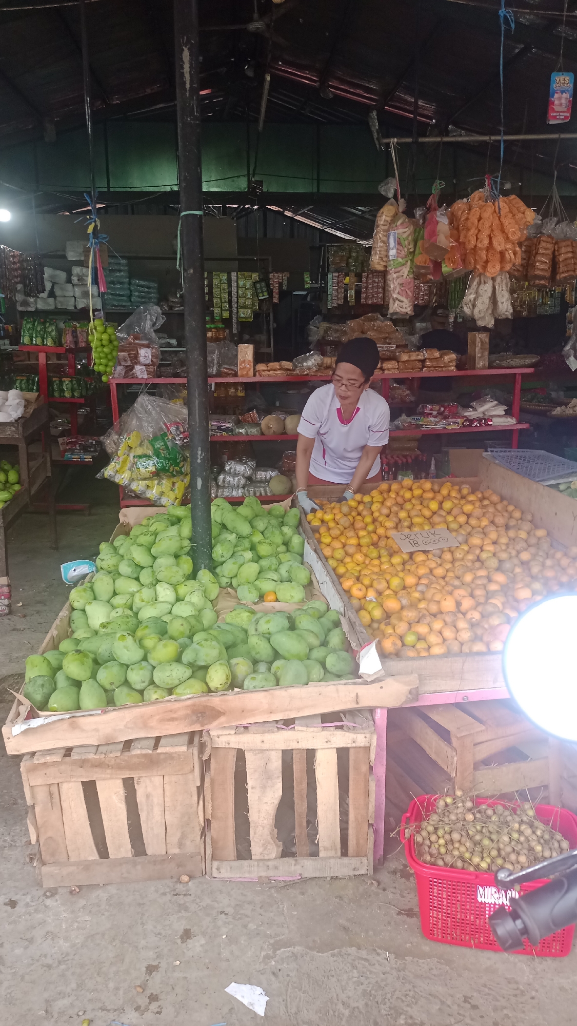 The image depicts an outdoor market stall in Karyamandala, Indonesia, filled with fresh produce. A woman, likely a vendor, stands behind a display of green mangoes and oranges. She is wearing glasses and a white shirt with pink trim. The fruit is arranged in large wooden crates. Behind the fruit, shelves are stocked with various packaged goods, including snacks and drinks. Hanging from the stall's roof are additional items, such as bags of snacks and what appear to be dried goods. In the foreground, a red basket holds small, round fruits, possibly longans. The ground is a concrete surface, indicating a public market setting. The overall scene suggests a bustling day at the local marketplace.