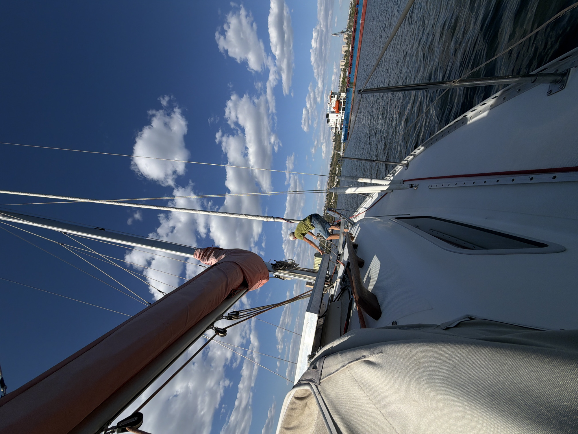 A person in a green t-shirt and blue shorts is standing on the deck of a white sailboat, leaning over and pulling a rope. The sailboat is on the water, with a large pink sail furled around the boom. In the distance, a large blue and red cargo ship is visible, as well as a crane on the shore. The sky is a clear blue with scattered white clouds. The location is the Volga River in Kamyshin, Russia.