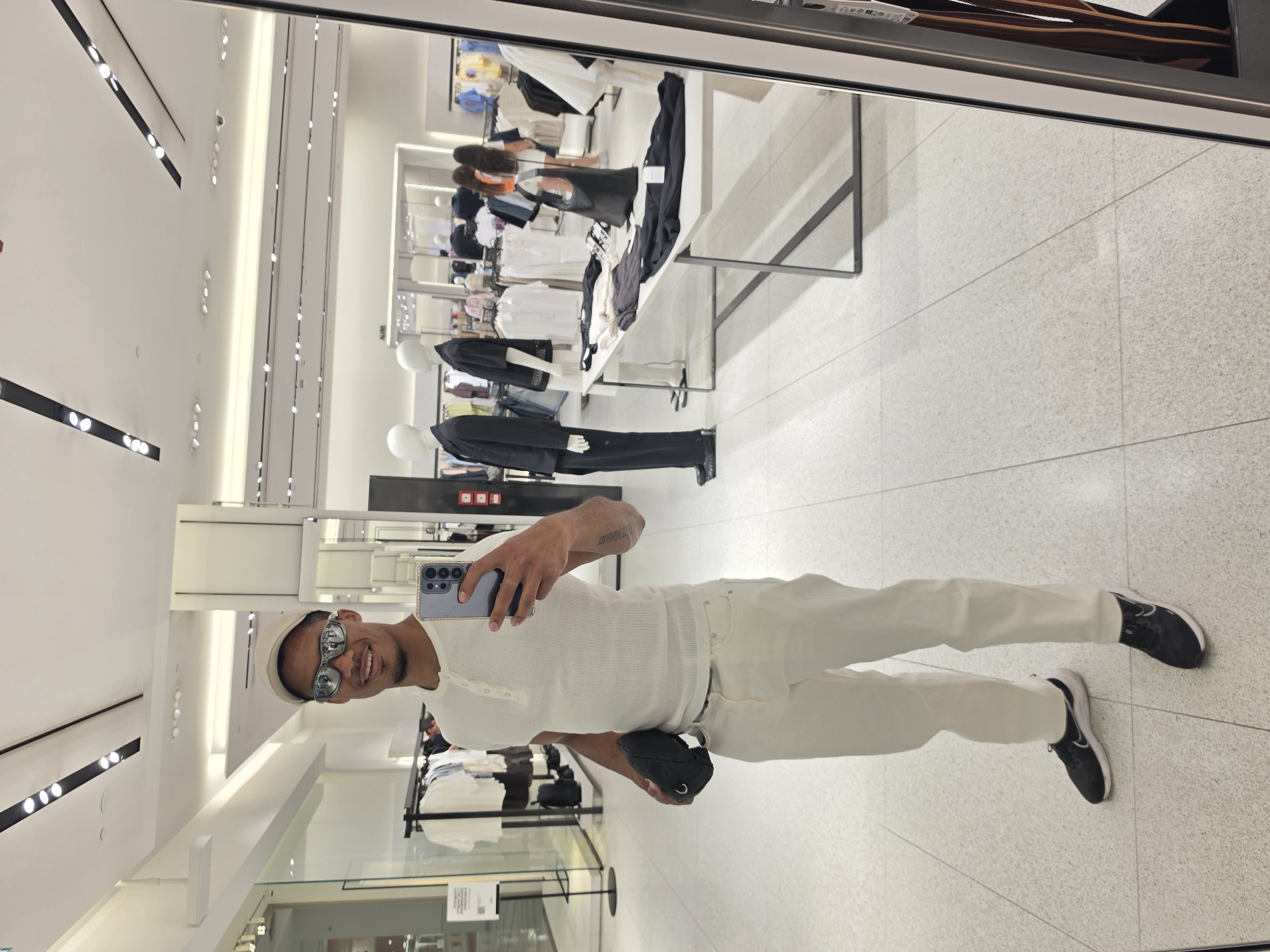 A young Black man stands in a brightly lit clothing store, taking a selfie in a mirror. He is wearing a white short-sleeved henley shirt, cream-colored pants, black sneakers, and a light-colored baseball cap. He has sunglasses on and is holding a black pouch in his left hand. The store appears to be a modern retail environment with white tiled floors, white walls, and track lighting. Mannequins dressed in clothing are visible in the background, along with racks of apparel and display tables. The store is clean and organized, with ample space for shoppers. The man's pose is casual and confident. The image captures a moment of personal style and shopping in a commercial setting.