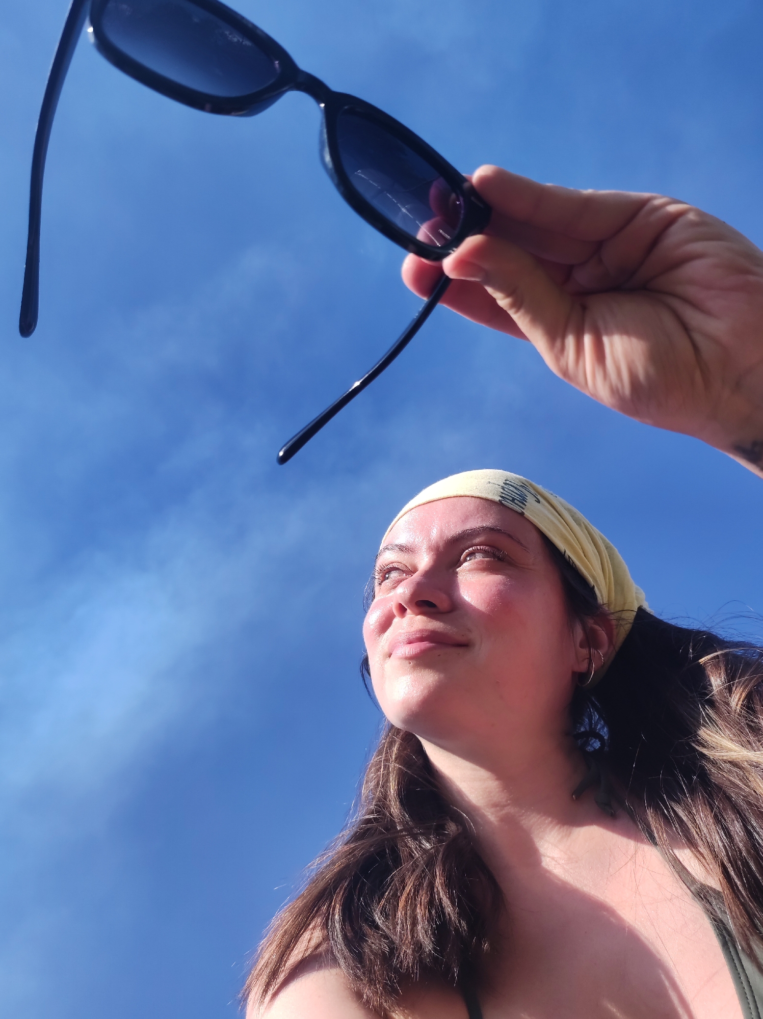 A low-angle shot captures a woman under a bright blue sky. She is wearing a pale yellow patterned bandana and earrings. Her brown hair flows over her shoulder. She is gazing upwards with a serene expression. Above her, a hand holds a pair of black sunglasses. The location is an outdoor setting, but the information does not give enough detail to precisely determine a location in Фоки, Russia.