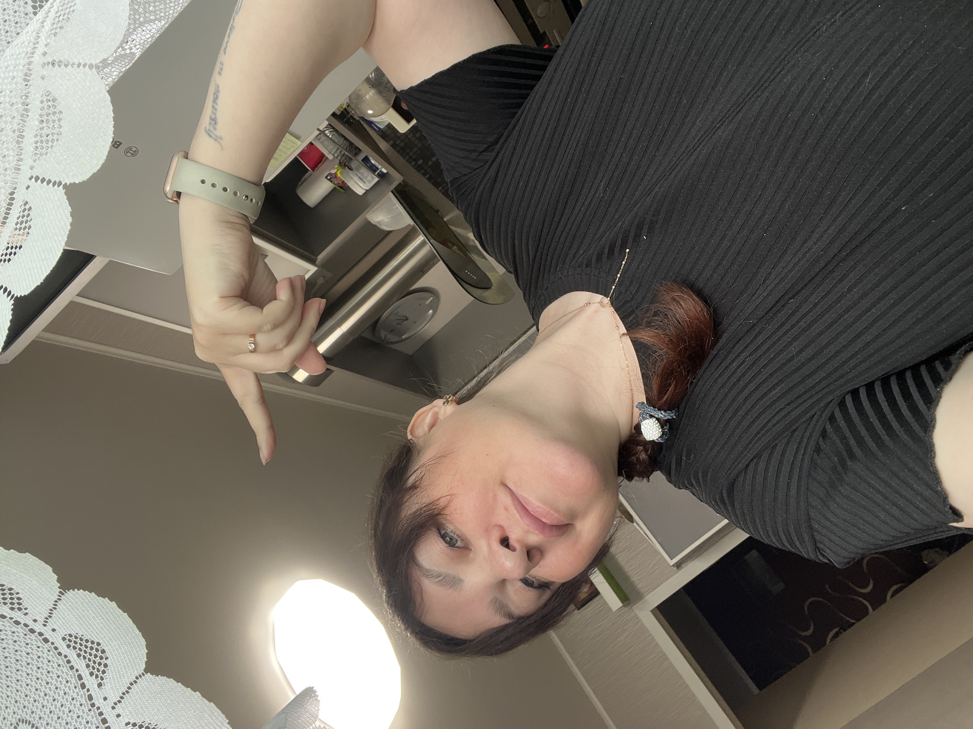 This image is a selfie of a woman taken from a low angle, with her looking downwards and slightly smiling at the camera. She has dark hair, pulled back, and wears a black ribbed top. On her left wrist, she has a light green/grey smartwatch with a rose gold-colored casing. A small tattoo reading "believe in yourself" is visible on her inner left forearm, and a gold-toned ring with a clear stone is on her left ring finger. A thin gold necklace and a decorative hair tie/clip with white and black details are also visible near her neck. Her left arm is raised and bent, with her index finger pointing downwards towards the camera.
The background suggests an indoor setting, likely a kitchen or a room with kitchen elements. To the left of the frame, a white lace fabric, possibly a curtain or tablecloth, is prominent. In the upper background, there are light-colored cabinets or appliances, including one with a visible "B" logo (possibly Bosch). Below this, stainless steel surfaces, various bottles, and containers are present. A bright, circular light source is visible in the lower left portion of the image. The overall impression is casual and personal, consistent with a self-portrait.