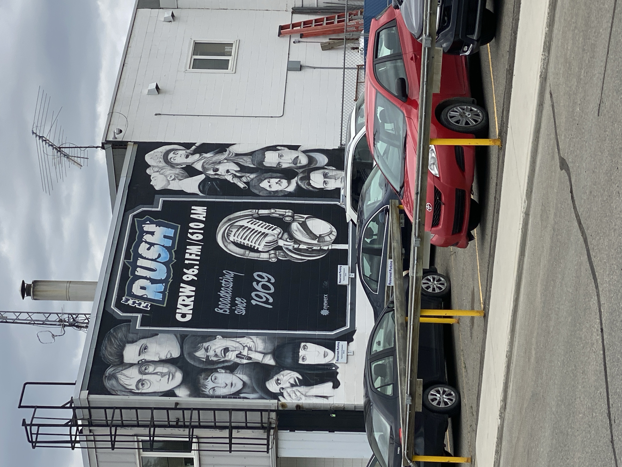 The image depicts a mural on the side of a building in Whitehorse, Canada, which features portraits of musicians and a vintage microphone with headphones. The mural is for "The Rush CKRW 96.1FM / 610 AM," indicating it's likely the exterior of a radio station. The text below reads "Broadcasting since 1969." Several parked cars, including a red Toyota and a black sedan, are visible in the foreground, with a yellow barrier separating the parking lot from a walkway. The building appears to be a commercial structure with a ladder attached to its side. The sky is overcast.