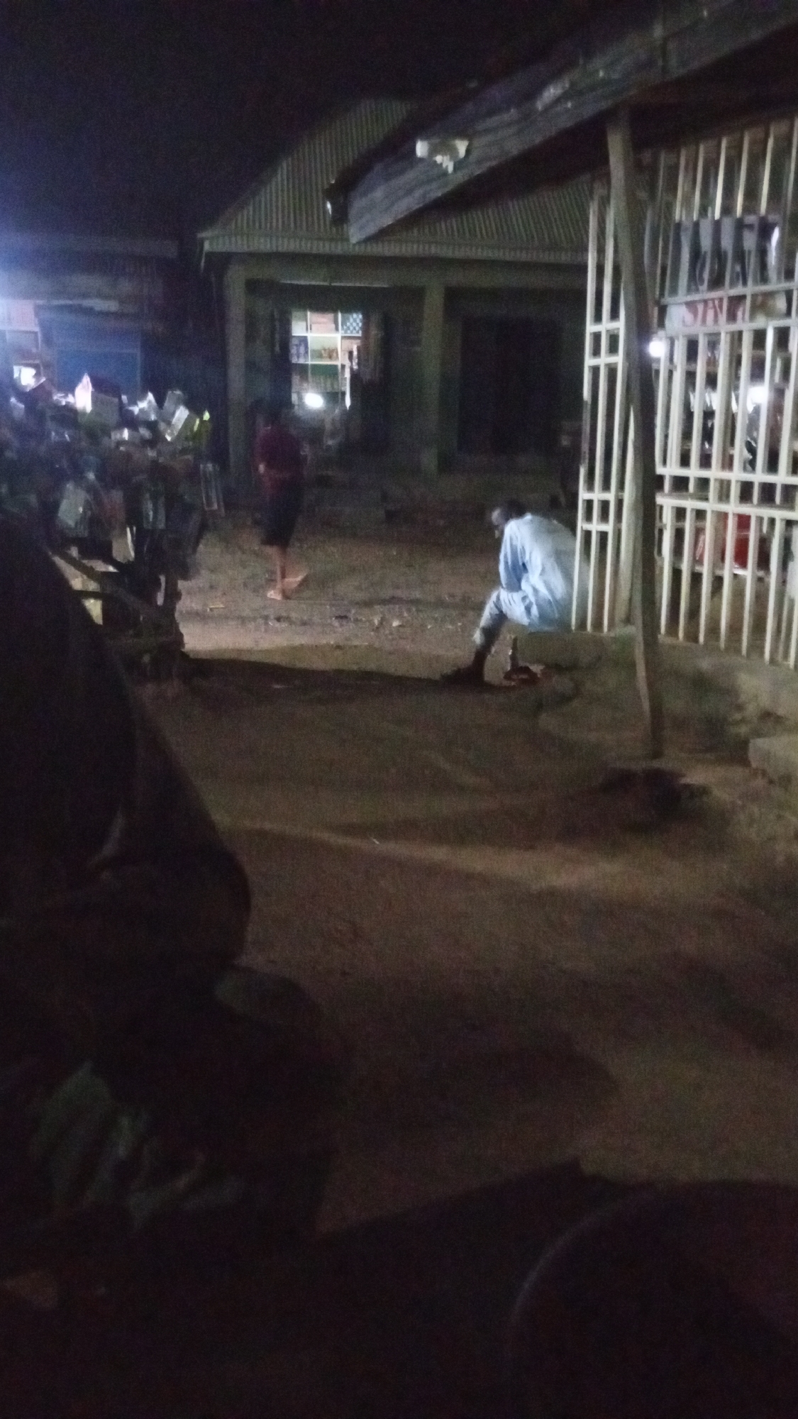 This night-time image, taken in Adankolo, Nigeria, captures a quiet street or market scene. The predominant darkness suggests late evening or night, with light sources primarily originating from within buildings or distant streetlights.
In the right foreground, a person clad in a light-colored, long-sleeved garment (possibly white or light blue) and dark lower attire is seated on a low concrete ledge or barrier. They are positioned against a tall white metal gate or fence and appear to be looking downwards, perhaps resting or in thought. A small, dark object, possibly a bottle, lies on the ground near their feet.
Towards the center of the frame, another individual, wearing a dark red top and dark shorts or trousers, is seen walking away from the viewer towards the left. They appear to be barefoot or wearing light-colored sandals.
The left side of the image features a partially obscured figure in the extreme foreground, largely covered by shadow. Further into the mid-ground on the left, a collection of objects, possibly merchandise or goods from a stall, are stacked and reflect light, suggesting they might be electronic items or products with reflective packaging. These are situated in front of a building with a corrugated metal roof.
More buildings with similar corrugated metal roofs are visible in the background, contributing to the setting of a local commercial area. The ground surface throughout the visible area is unpaved, appearing to be dirt or gravel. The atmosphere is calm, with limited activity. No discernible text is visible anywhere in the image.