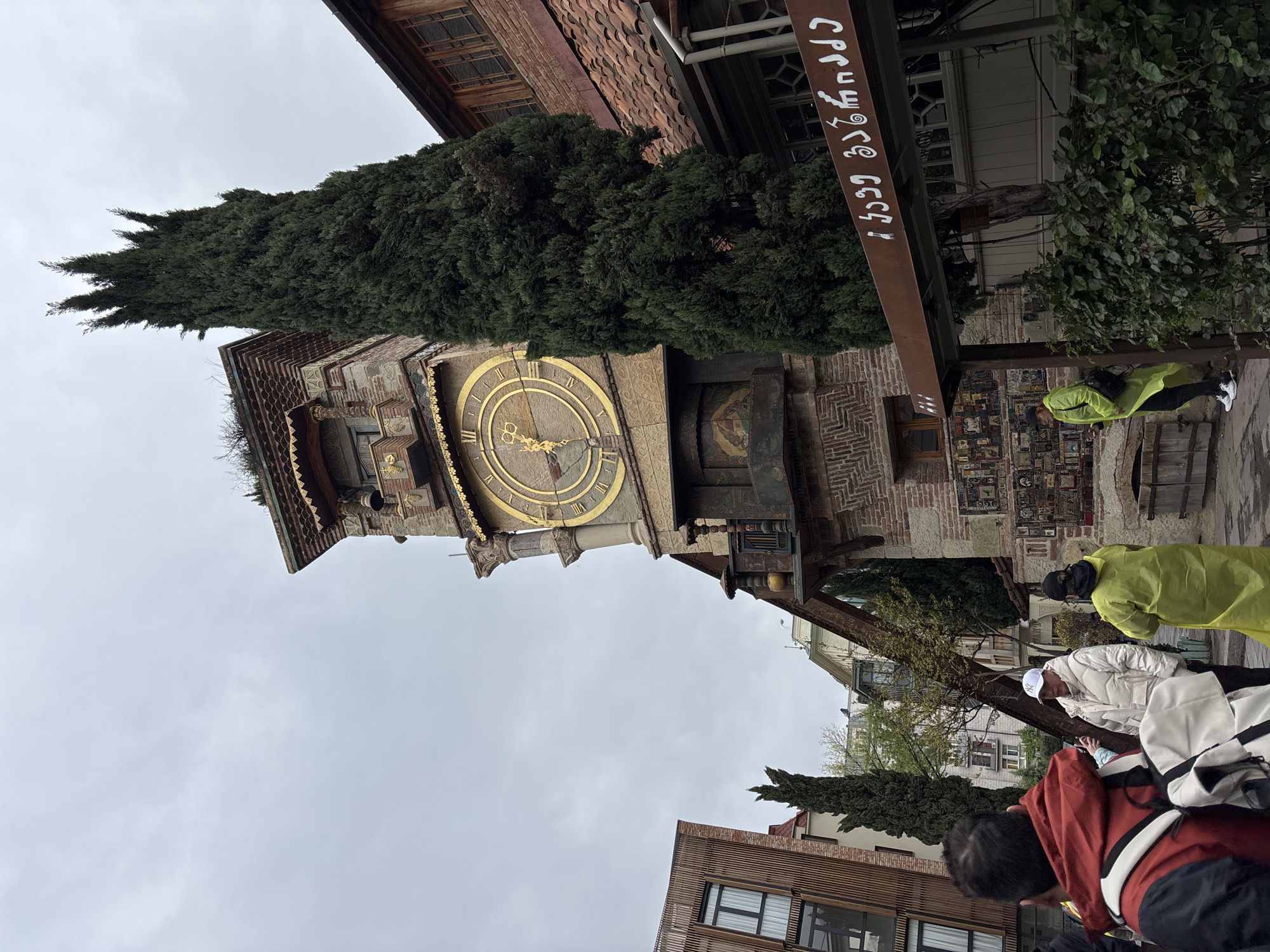 The image shows a tilted view of the Rezo Gabriadze Clock Tower in Tbilisi, Georgia. The ornate clock tower, with its visible golden clock face, is flanked by a tall, thin tree. A metal sign with Georgian script hangs near the tower. Below, several individuals are visible, including one in a bright yellow raincoat. The surrounding buildings are brick and modern in style. The sky is overcast.