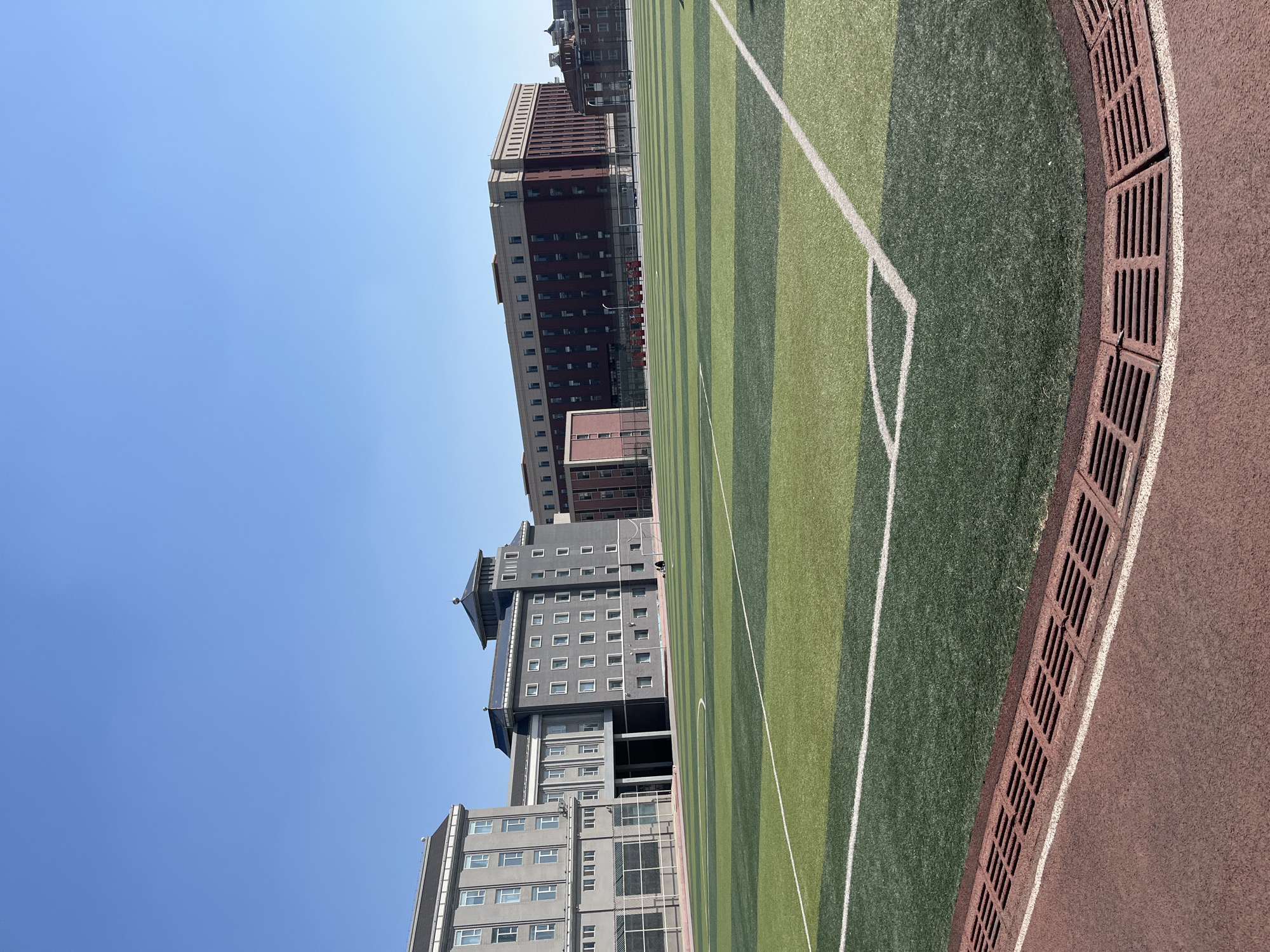 The image shows an outdoor scene in Changchun City, China, featuring a soccer field with alternating stripes of light and dark green artificial turf. White lines mark the boundaries of the field. Adjacent to the field is a running track with a brown surface, separated from the turf by a drainage system with a brown grate.
Several multi-story buildings stand in the background. These structures vary in height and design, with one building being a red brick color. The sky above is clear and blue.