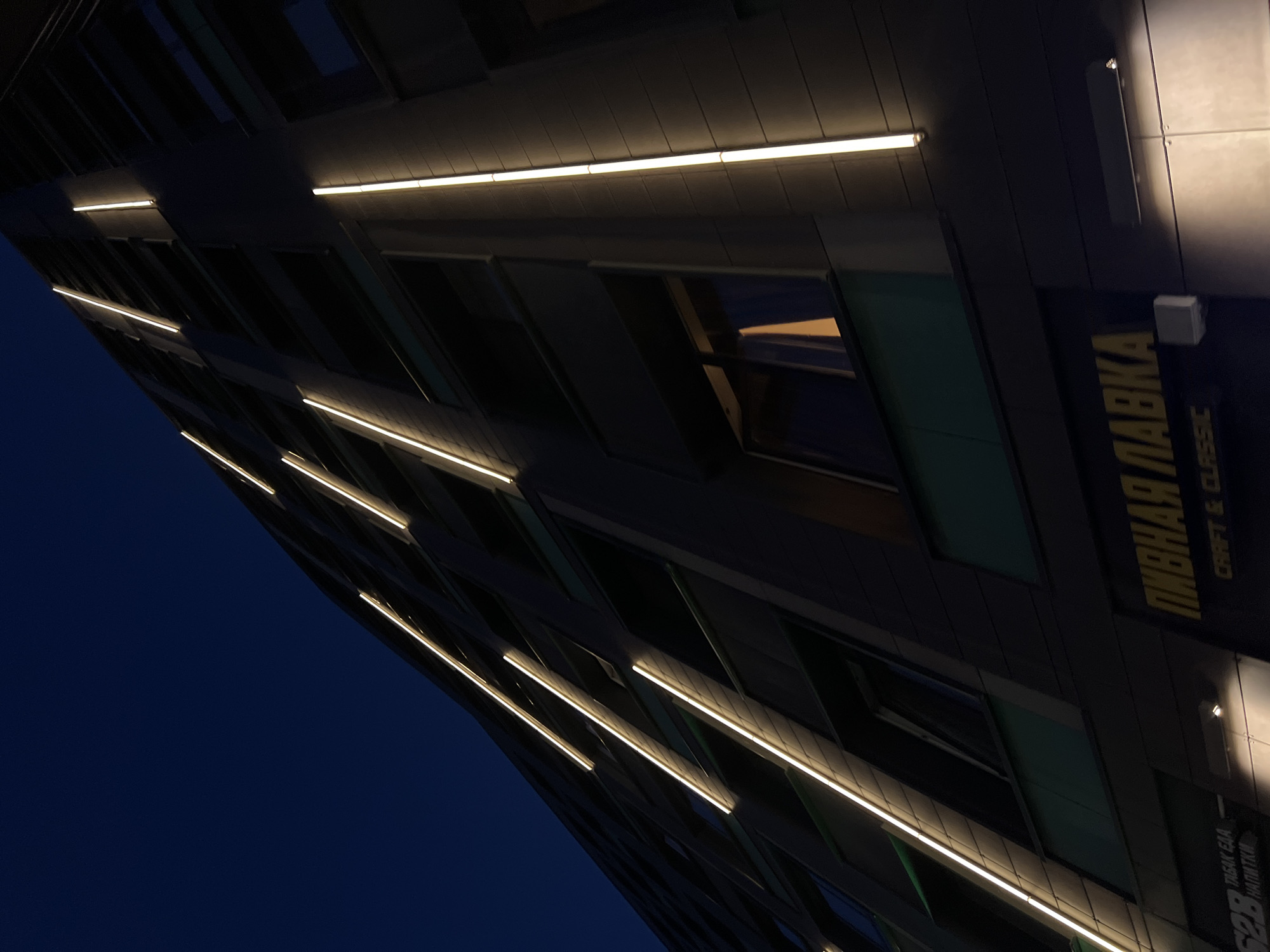 The image shows a modern building at night in Russia. The building has a dark facade with vertical lines of warm white LED lights running up its exterior. There are windows on several floors, and some are illuminated from within. In the lower part of the image, there is signage for a "Pivnaya Lavka" (Beer Shop) with the text "CRAFT & CLASSIC" below it. To the left of this sign, there is another partially visible sign that reads "TABAK EDA NAPITKI" (Tobacco Food Drinks). The sky is a deep, dark blue, indicating it is nighttime. The perspective is looking up at the building from a low angle.
