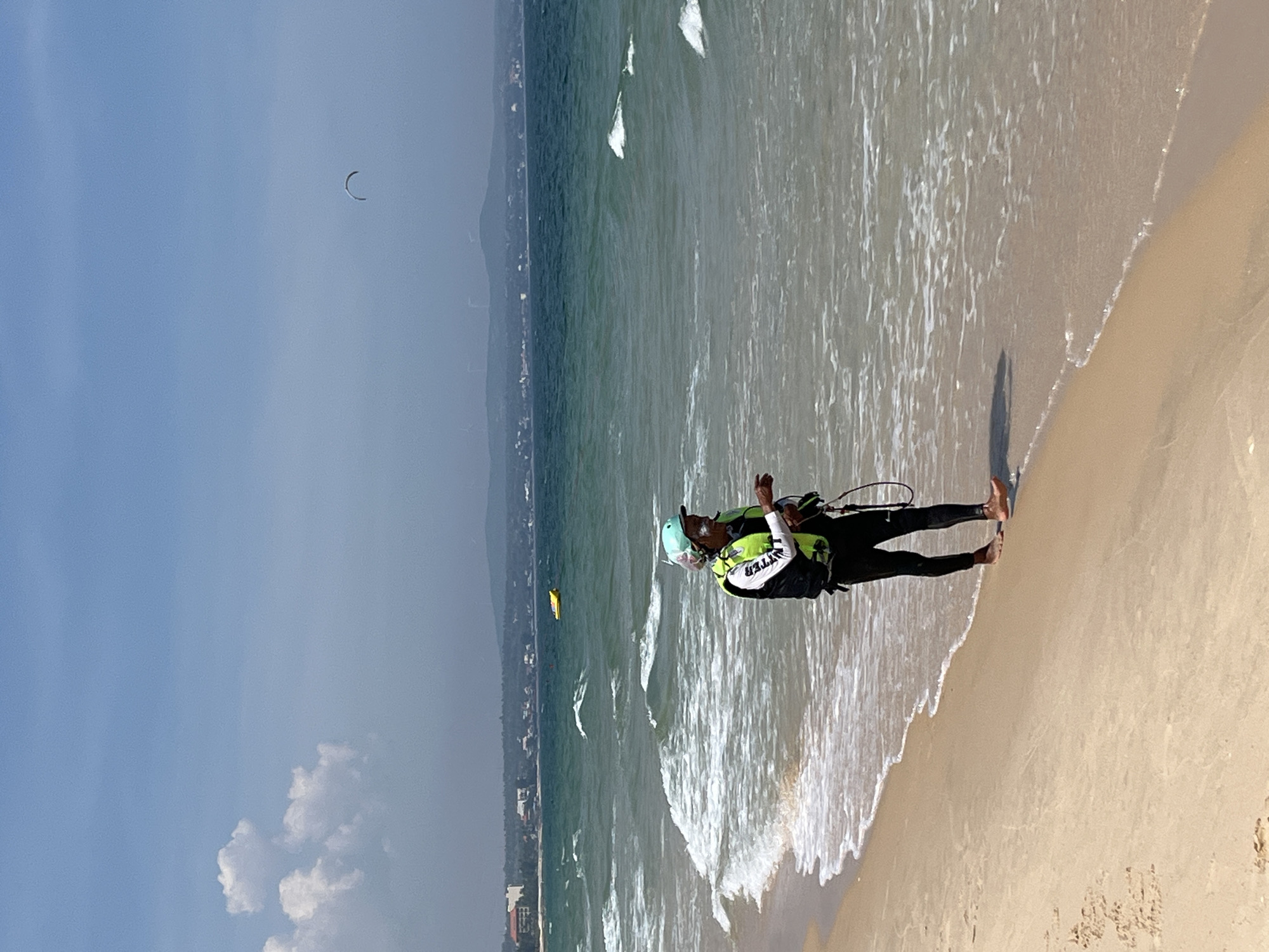 A person stands on a sandy beach at the edge of the turquoise ocean in Phan Thiet, Vietnam. The person is wearing a black wetsuit, a bright green life vest, and a light blue helmet. They are holding a kiteboarding bar and lines. The ocean has moderate waves with white foam breaking near the shore. In the background, a yellow float is visible in the water, and beyond that, a coastline with buildings and hills. The sky is mostly clear blue with a few scattered clouds. A kiteboarding kite is visible in the far distance. The scene appears to be set on a sunny day, likely during daylight hours.