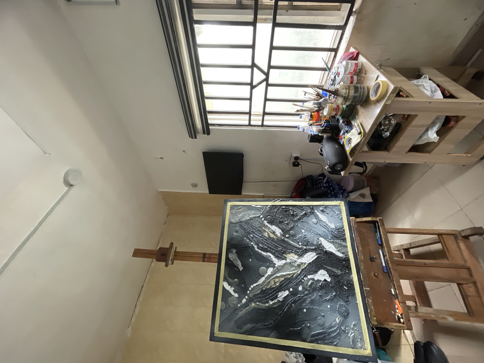 The image shows an interior space likely in Ado, Nigeria, given the prompt. An easel stands holding a square canvas with an abstract painting predominantly in black, white, and gold. The painting features swirling patterns. To the right of the easel is a wooden table filled with paint bottles, brushes, and other artist supplies. A window with a black grid frame is above the table, providing natural light. Below the window hangs a small, dark television. Bags and personal items are scattered on the floor near the table. The room's walls are painted in a light color. No people are visible in the shot.