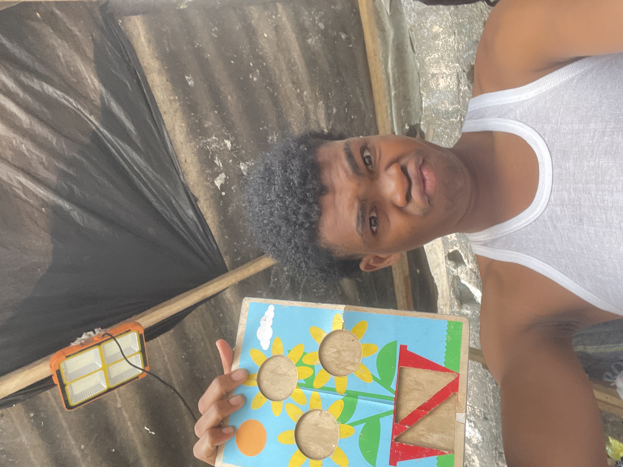 A young Black male is holding up a wooden puzzle board in front of him. The puzzle board is decorated with a blue sky, a yellow sun, three yellow sunflowers with circular cutouts, green stems, and a red and green flower pot. The young male is wearing a white tank top and has dark, curly hair. He is looking at the camera with a playful expression. Behind him, the scene appears to be outdoors, possibly in a construction or workshop area. A corrugated metal roof is visible above, with a black tarp draped over it. An orange and white solar-powered light fixture is attached to the roof. The wall behind the male is rough and textured, suggesting it is made of concrete or stone. The ground is also visible, appearing to be made of dirt and debris. The overall setting suggests a casual, informal environment, possibly in Ilado, Nigeria, given the rustic and utilitarian nature of the surroundings.
