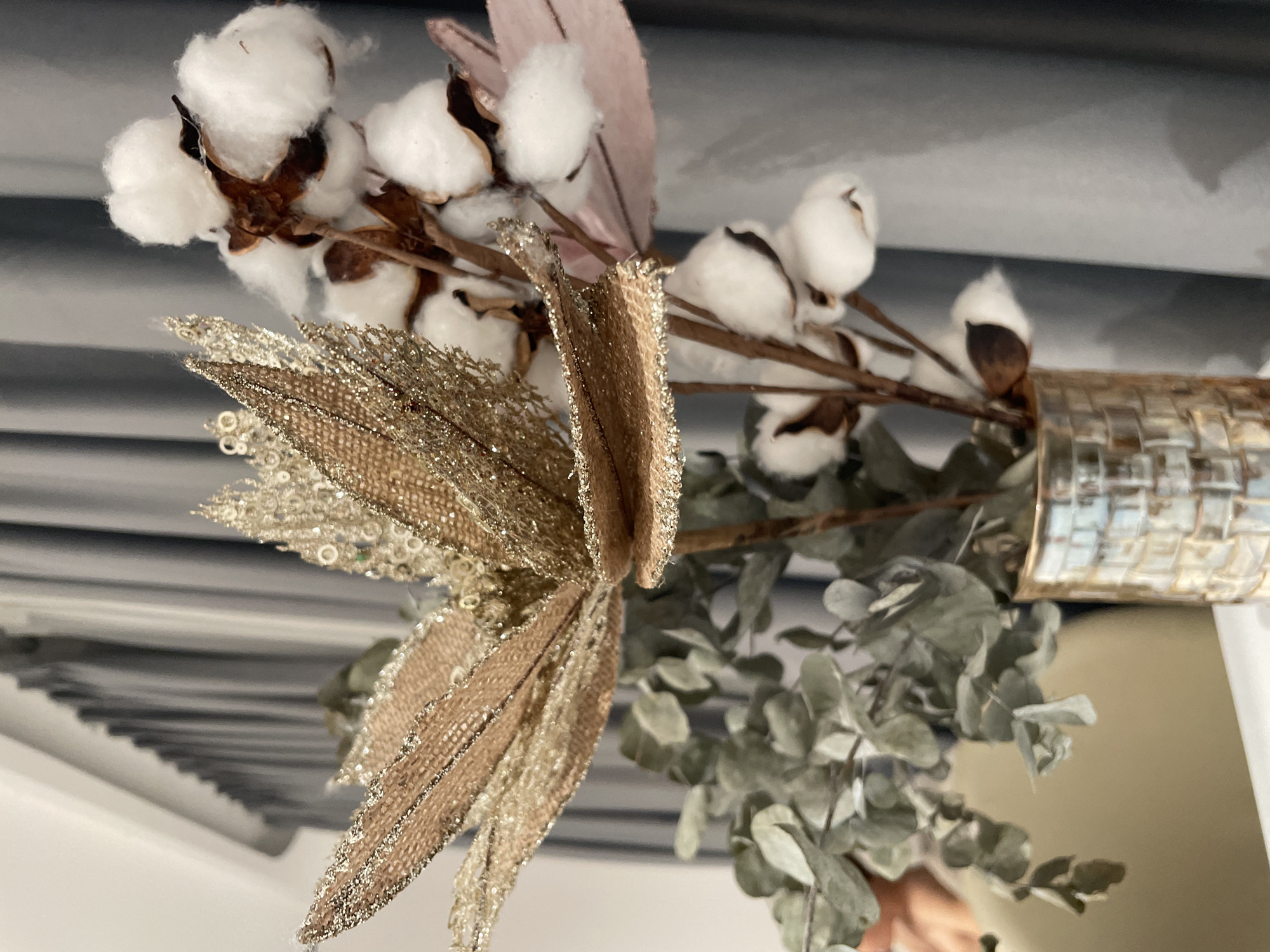 This close-up image depicts a decorative arrangement, possibly a centerpiece or wall decor, set in an indoor environment in Bắc Nha Trang, Vietnam. The main subjects are several branches featuring fluffy white cotton bolls, each emerging from a dark brown husk. Interspersed among the cotton are artificial decorative elements, including prominent gold, sparkly faux foliage or ribbon, which add a festive or glamorous touch. Beneath these, dried eucalyptus leaves with a muted grayish-green color are visible, contributing a natural element to the composition. The arrangement is held in what appears to be a textured, possibly golden or metallic, cylindrical container or vase, a small portion of which is visible on the right. The background features horizontal dark lines against a lighter surface, suggesting a textured wall, blinds, or corrugated material. No activity or event is taking place, as it is a still life. The scene conveys a sense of warmth and decorative elegance. There is no visible text.