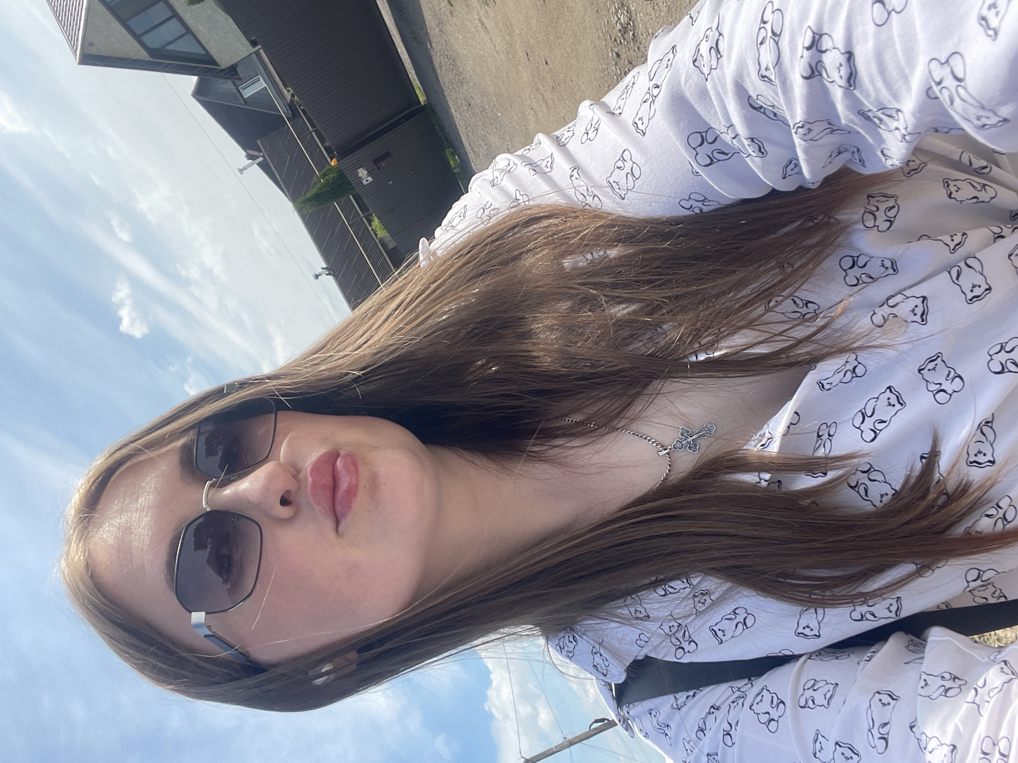 A close-up, low-angle selfie-style photograph depicts an individual, likely female, with long, straight brown hair, outdoors in Сырково, Russia. She wears dark, hexagonal sunglasses with a reflective surface, and her lips are slightly puckered with a glossy finish. A silver-colored chain with a cross pendant hangs around her neck. She is wearing a white long-sleeved shirt patterned with black outline gummy bear figures. The background features a clear blue sky with sparse white clouds. Below the sky, several residential buildings with dark-tiled roofs are visible. A light-colored house with multiple windows, including an arched one, stands prominently on the far right. A dark fence or gate, possibly open, separates the foreground from the houses, with a slender conical evergreen tree positioned behind it. A dirt or gravel path is visible in the lower right portion of the frame.