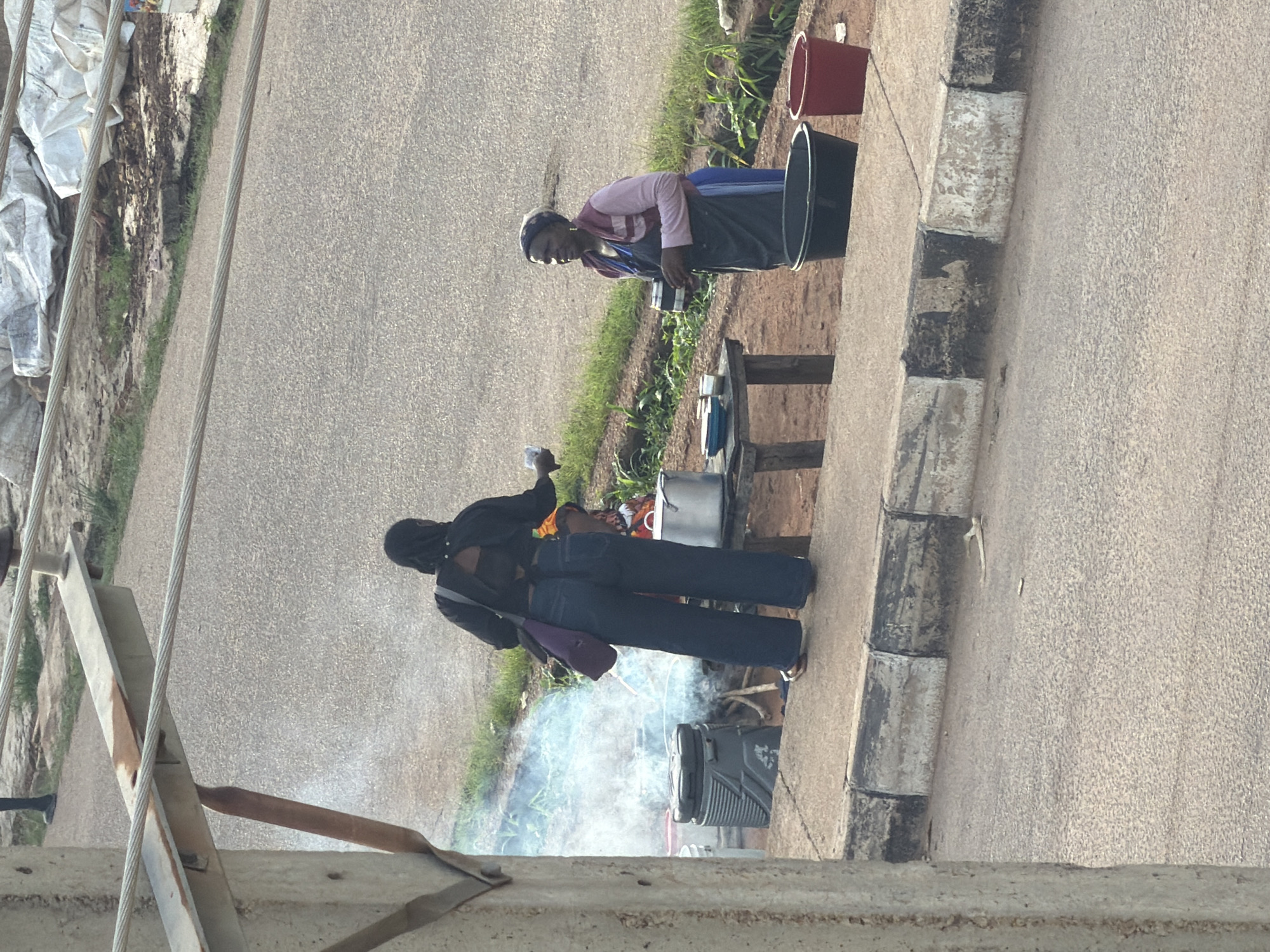 Two women are standing on a paved roadside in what is described as Gura Top, Nigeria. The woman on the left is wearing a dark headscarf, a black top, and dark blue jeans, and is holding a cup in her right hand. She is facing away from the camera, looking towards the woman on the right. She has a purple bag slung over her left shoulder. The woman on the right is wearing a headscarf, a pink and red top, and a dark blue apron. She is holding a cup in her left hand and facing towards the woman on the left. In the background, there is a large metal pot on a wooden table, and a grey cooler with a black lid. Smoke is rising from a fire under the pot. There are two large buckets, one black and one red, on the sidewalk to the right of the women. The ground is a mix of asphalt and dirt, with some green grass and vegetation visible. There are also some white tarps or bags piled up in the background.