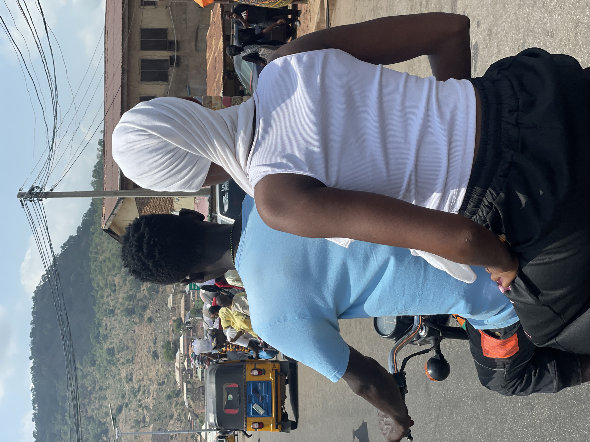 Two individuals are on a motorcycle navigating a busy street in Lokoja, Nigeria. The foreground shows the back view of the motorcycle rider, wearing a light blue t-shirt and black pants with orange accents. The passenger behind the driver is wearing a white ribbed top and a white headscarf. The street is congested with pedestrians and vehicles, including a yellow auto rickshaw (keke) with visible registration details. A mountain rises in the background. Overhead power lines stretch across the sky. Buildings with corrugated metal roofs are visible along the roadside.