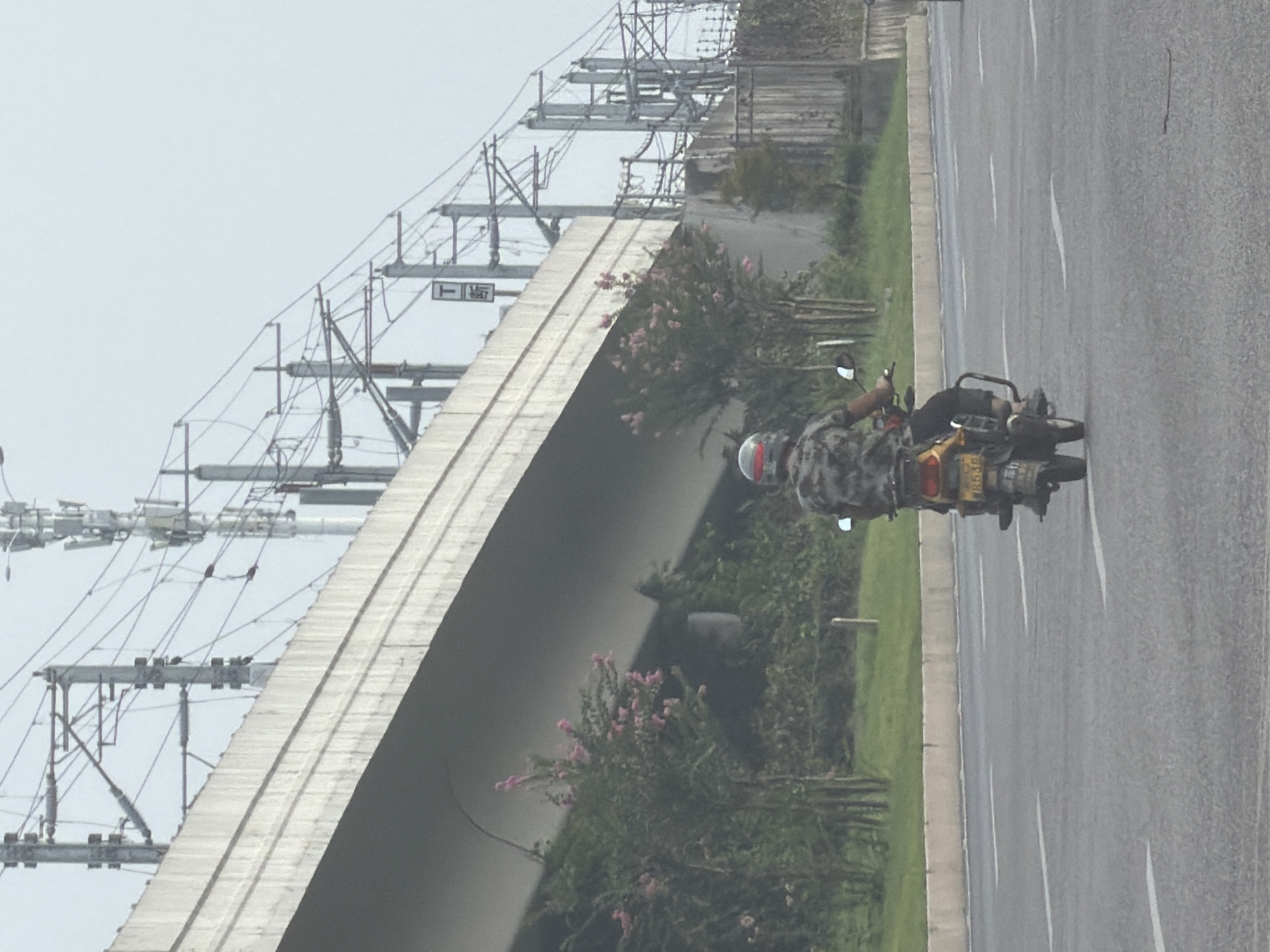 The image shows a person riding a motorcycle on a road in Jiangjin, China. The motorcycle rider is wearing a camouflage shirt and a helmet. The road is paved and has white lines marking the lanes. In the background, there is an elevated concrete structure, possibly a bridge or overpass, with power lines and utility poles. Trees and some vegetation are visible along the side of the road and under the elevated structure. The sky is overcast. The license plate on the motorcycle is partially visible and appears to have Chinese characters.