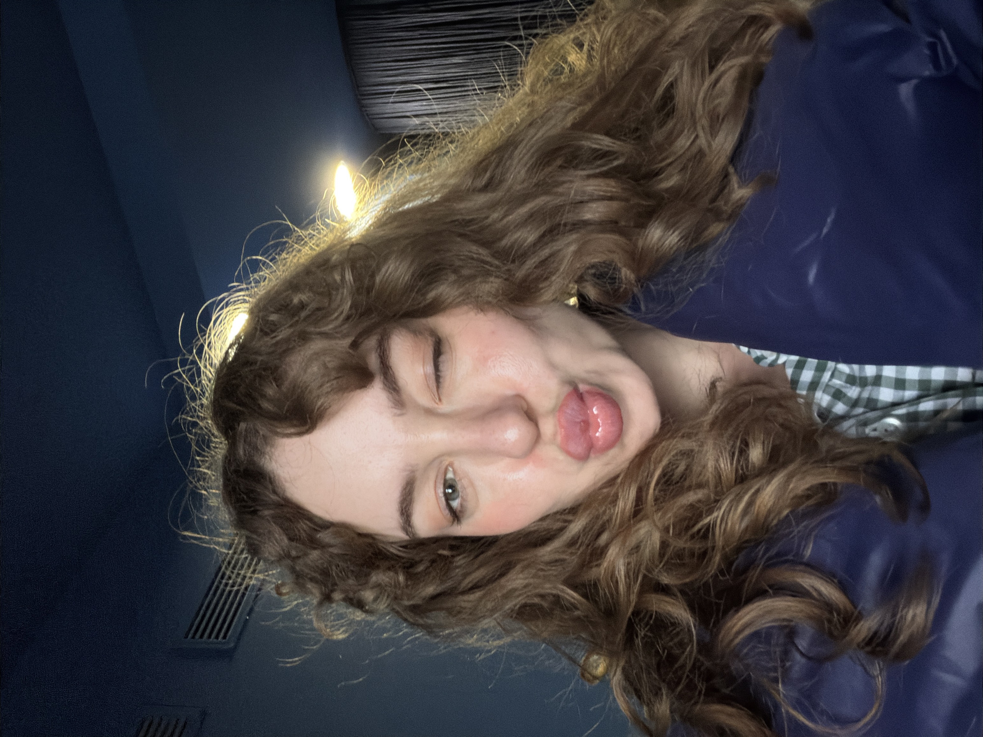 A female with long, curly brown hair is visible. She is winking and sticking her tongue out. She is wearing a blue garment and a checkered shirt. The background is dark blue. There is a vent in the wall. The location cannot be determined.