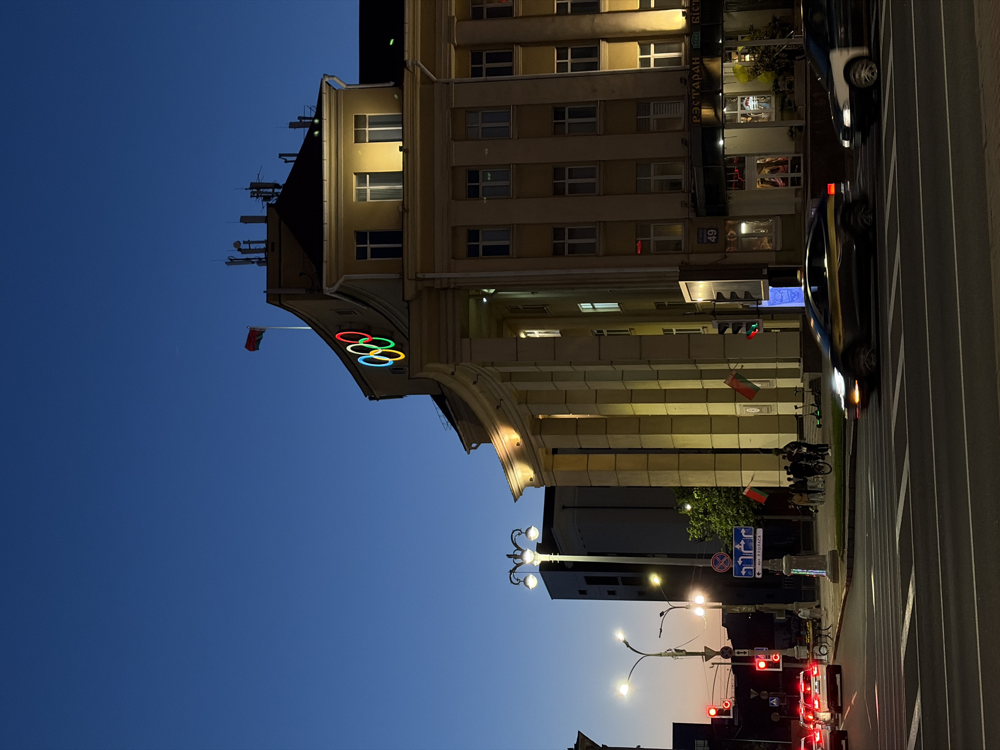 The image captures a street scene in Minsk, Belarus, at dusk. The sky transitions from deep blue to a lighter hue towards the horizon. Prominently featured is a building adorned with the Olympic rings logo and Belarusian flags, indicating it may be related to sports or government. Illuminated windows suggest activity inside the building. Street lights and vehicle headlights are visible, adding to the urban ambience. A crosswalk stretches across the street, and cars are present, indicating traffic. A group of people are standing near a street sign. The general impression is that of a bustling cityscape at twilight.