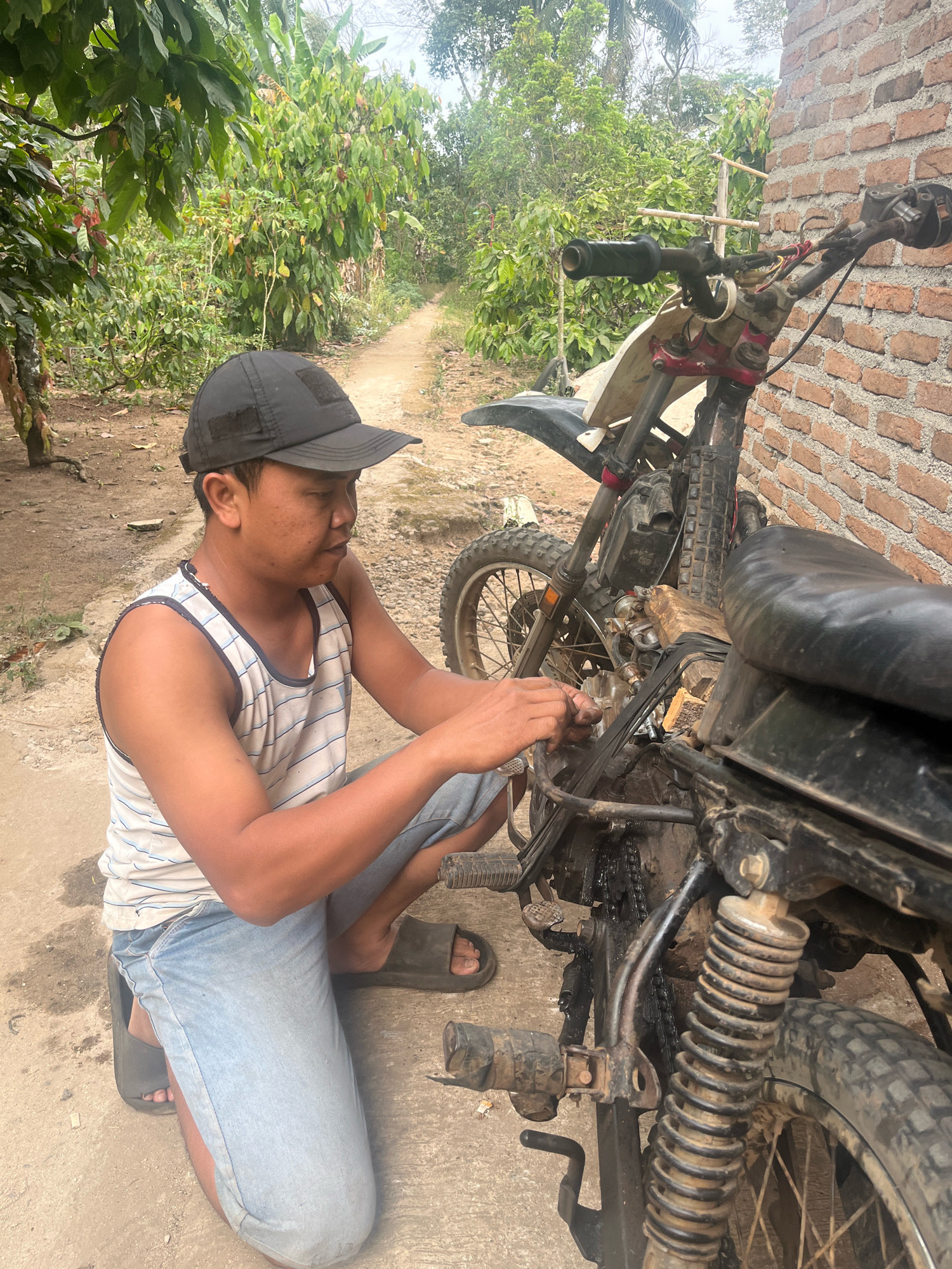 A man wearing a black baseball cap, a white and blue striped tank top, and light blue jeans is kneeling on a dirt path next to a dirt bike. He is wearing dark grey slides on his feet. The man appears to be working on the engine of the dirt bike, which is black and appears to be in a rural or unpaved area. The background consists of lush green vegetation, a dirt path, and a brick wall. The scene is consistent with a setting in Kebumen, Indonesia, given the typical rural landscape and architecture.