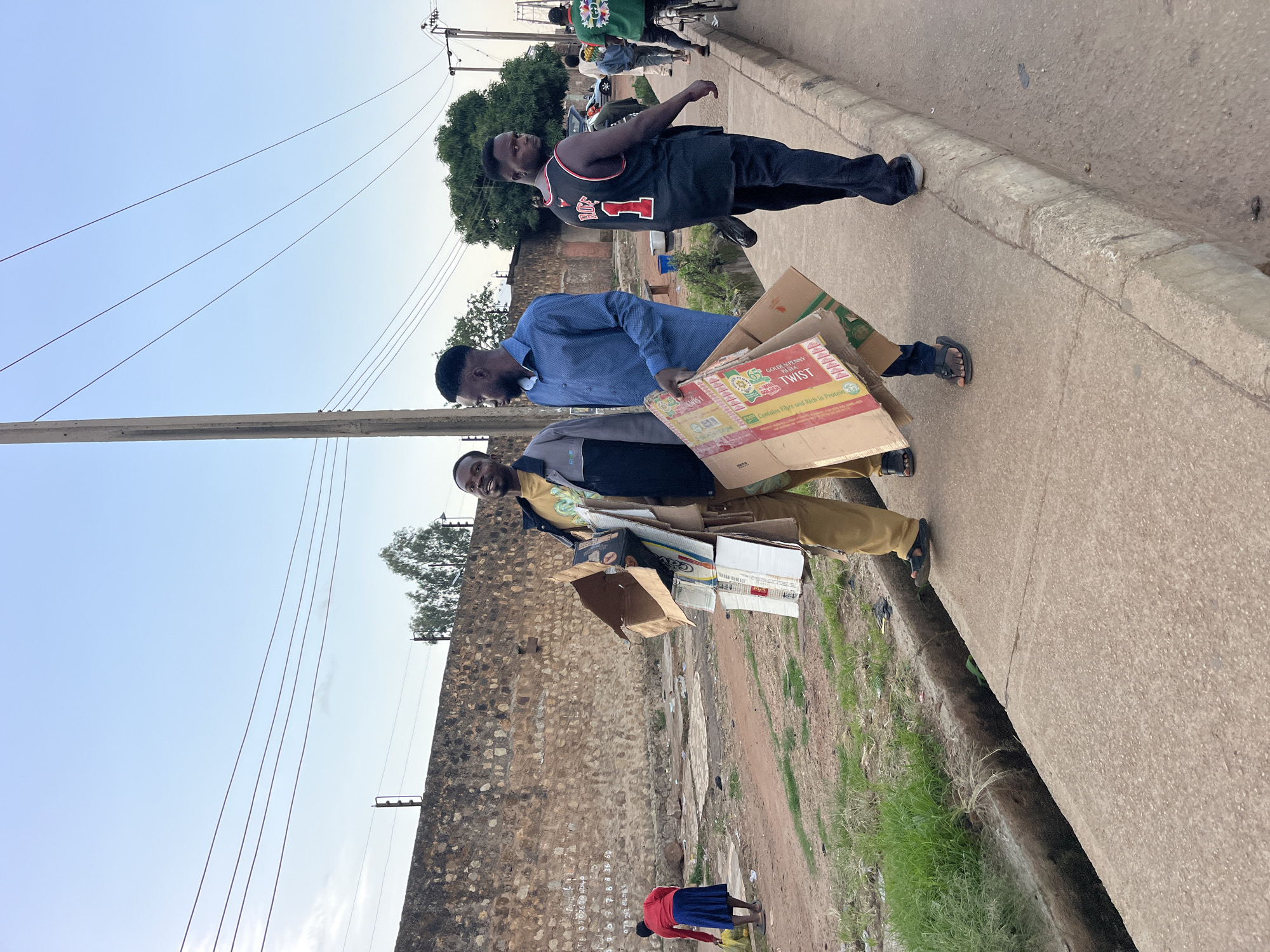 Three men are walking on a sidewalk in an urban area. The man in the foreground on the left is wearing a yellow t-shirt and tan pants, and is carrying a large stack of cardboard boxes. He is smiling. Next to him, a man in a blue shirt is walking and looking towards the man carrying the boxes. To the right, a man wearing a black tank top with the number "1" on it is walking away from the camera. In the background, there is a stone wall, some trees, and power lines. A woman in a red shirt and blue skirt is walking on a dirt path in the distance. The environment appears to be a street with a drainage ditch along the sidewalk.