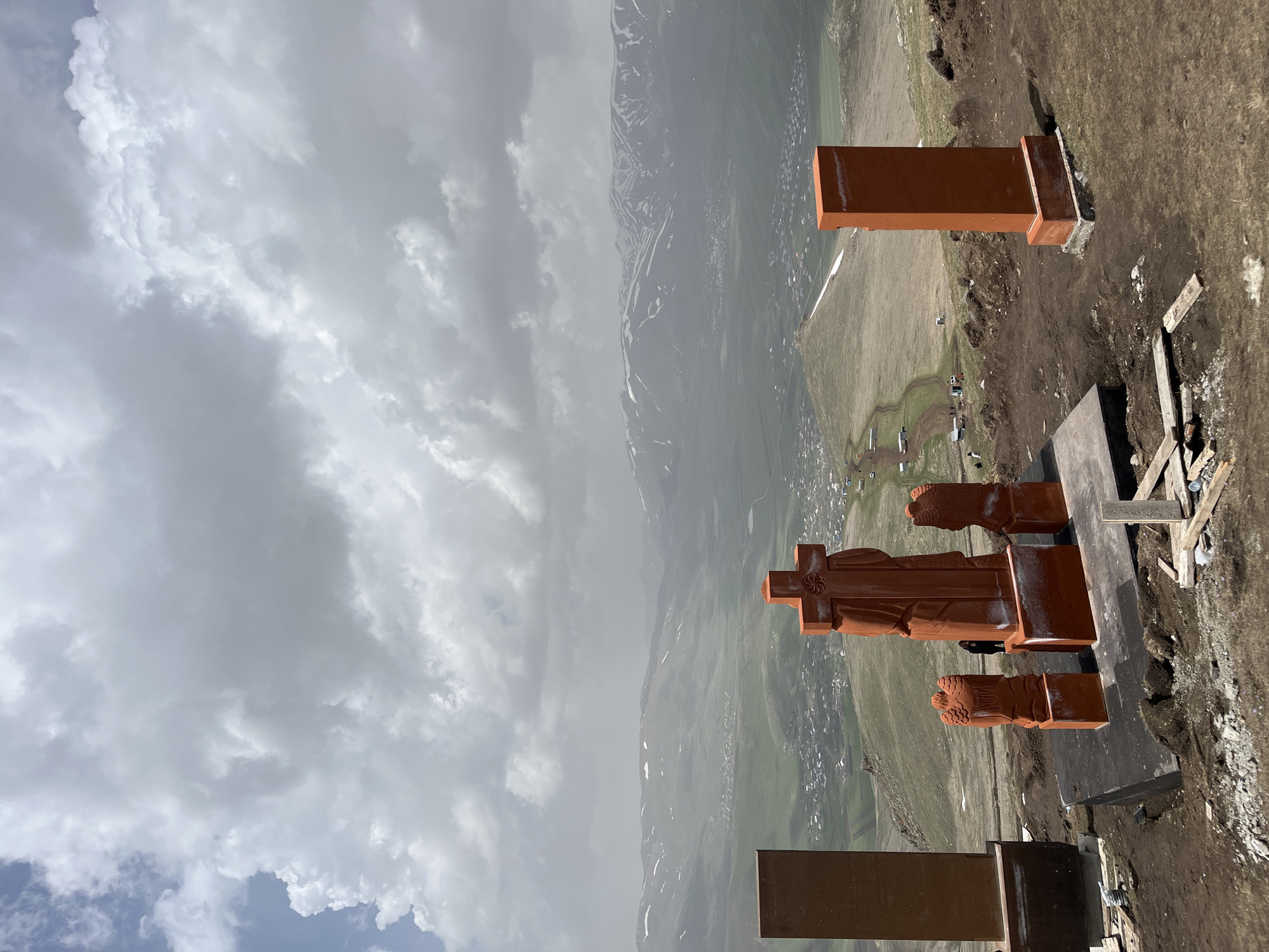 The image shows the Vardapet Memorial in Hartagyugh, Armenia. The Vardapet Memorial features a large, rust-colored cross flanked by stylized figures. The cross and figures are set upon a gray platform. Nearby are rectangular blocks, also rust-colored. The background shows a landscape with mountains and a sky filled with clouds. Buildings are visible in the valley below. The area around the memorial appears to be dirt and grass. A few pieces of wood debris are also visible.