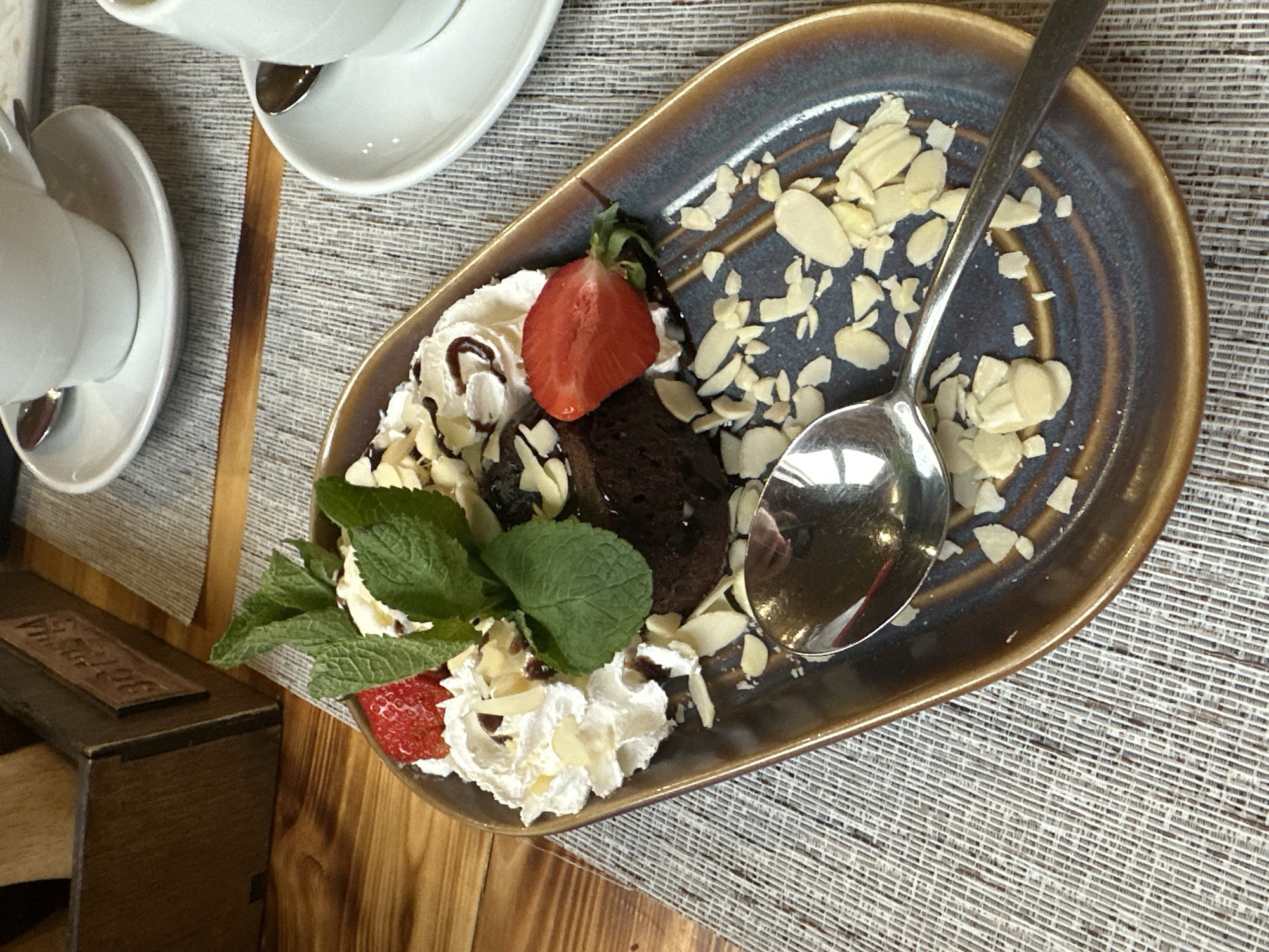 On a wooden table, a plate of dessert sits atop a woven placemat. The dessert consists of a dark brown cake, whipped cream, sliced strawberries, mint leaves, and sliced almonds. A silver spoon rests on the plate. To the left of the plate, a small wooden box is visible. Above the plate, two white teacups and saucers are positioned. The location is Unknown, Unknown.