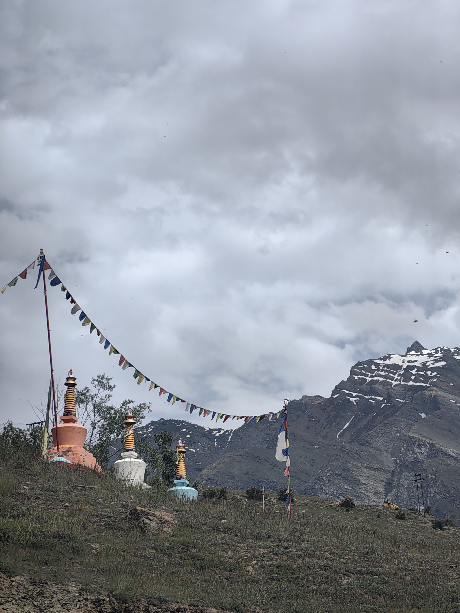 A medium shot captures a serene landscape in Tandi, India, under a cloudy sky. In the foreground, a grassy slope is dotted with three Tibetan stupas, or chortens. The stupas, arranged from left to right, are distinct in color and design. The leftmost stupa is large and orange, with a blue base. The middle stupa is white with golden ornamentation, and the rightmost stupa is light blue with golden accents. Prayer flags, in various colors, are strung across the frame, a long string of them extending from a pole on the left and curving towards the right. Behind the stupas, a snow-capped mountain range dominates the background, its rocky slopes contrasting with the white snow. The overall atmosphere is one of tranquility and spirituality.