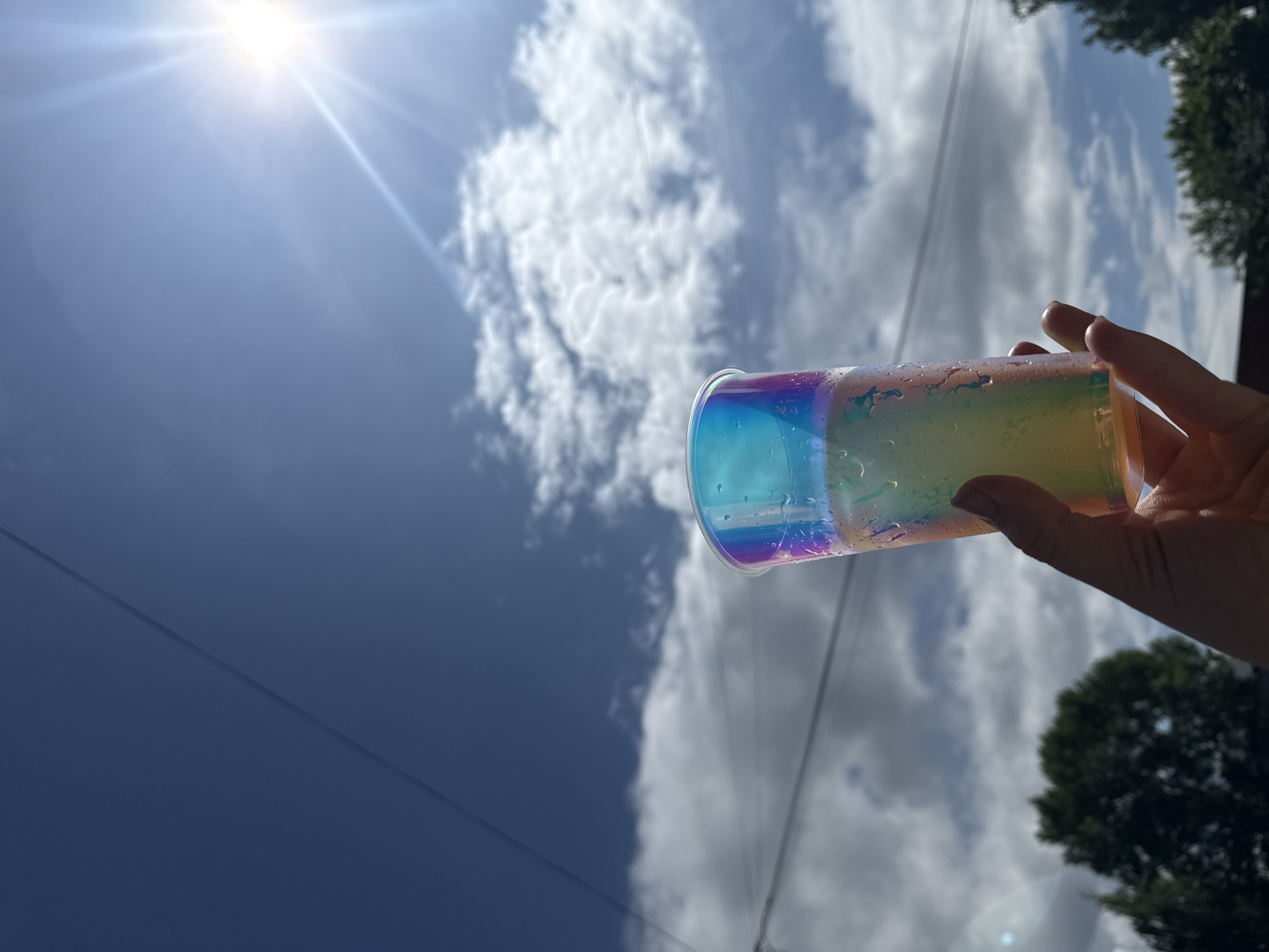 A low-angle shot captures a hand holding a clear, iridescent plastic cup filled with a pale yellow liquid. The cup, with condensation on its surface, is positioned in the foreground against a backdrop of a bright blue sky with scattered white clouds. The sun is visible as a bright, starburst-shaped lens flare in the upper right corner. A dark, thin line, possibly a power line, stretches diagonally across the sky. In the lower part of the frame, the tops of dark green trees are visible. The scene appears to be an outdoor setting, likely during the day. There is no discernible location or time indicator within the image itself that specifically places it in Chaikovsky, Russia.