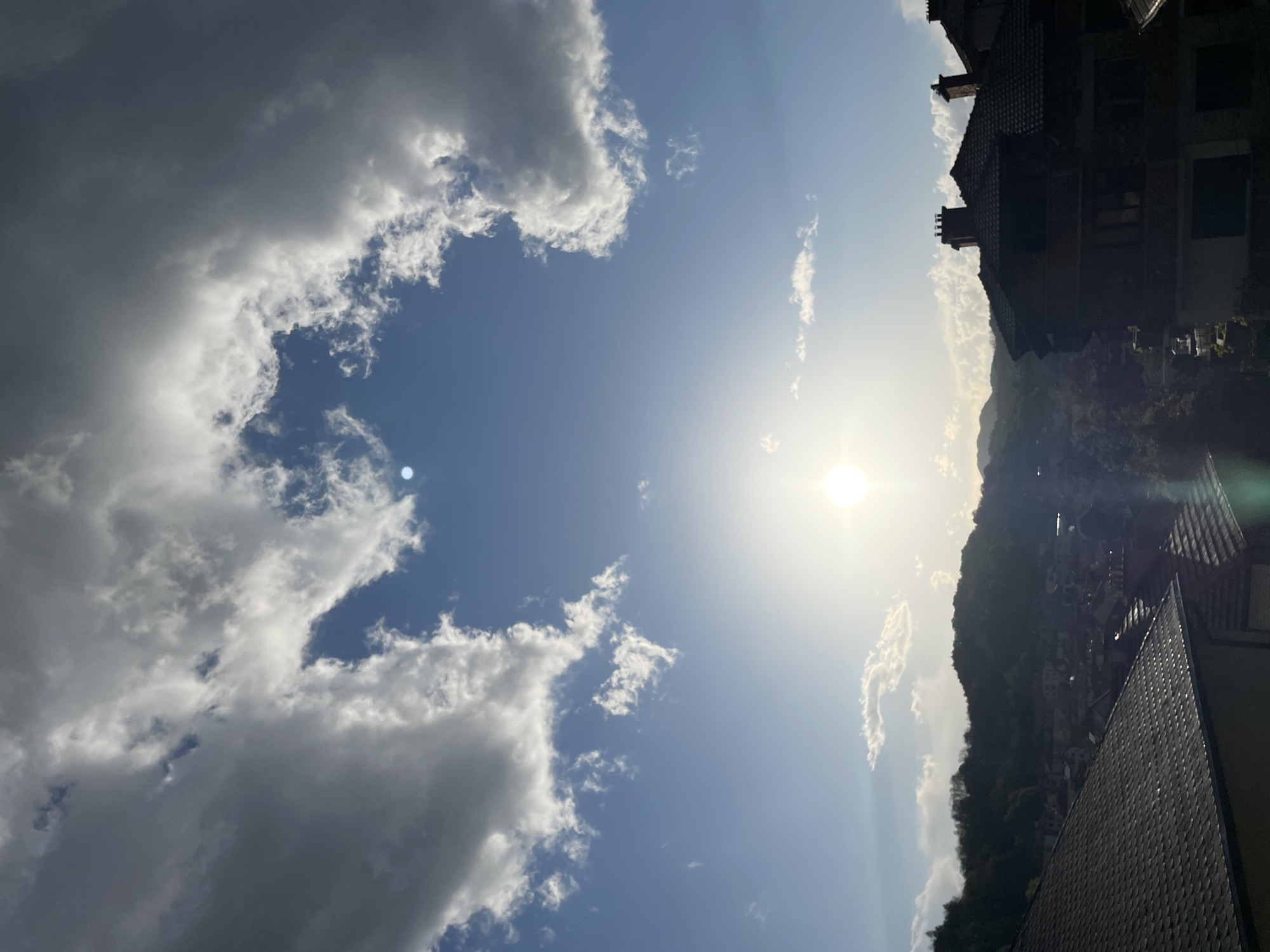The image shows a daytime scene of a sky with blue patches, intermittent white clouds, and the sun. The right side of the frame includes the outline of a hill covered in trees, the roof of a building in the foreground, and a section of buildings. The location is identified as Zlatograd, Bulgaria.