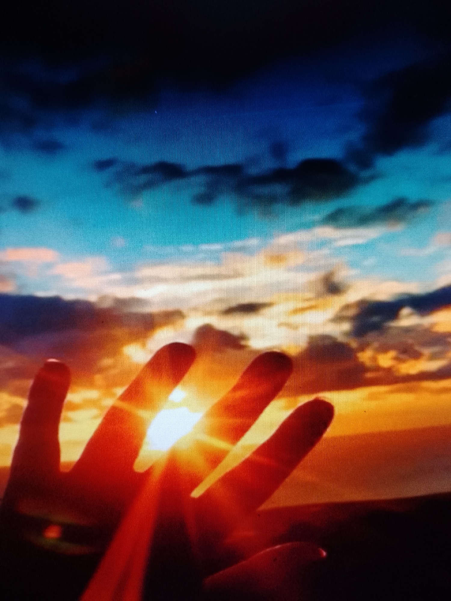 A hand is held up toward a bright orange and yellow sunset. The sky is also blue, with dark clouds. The location is Unknown, Unknown.