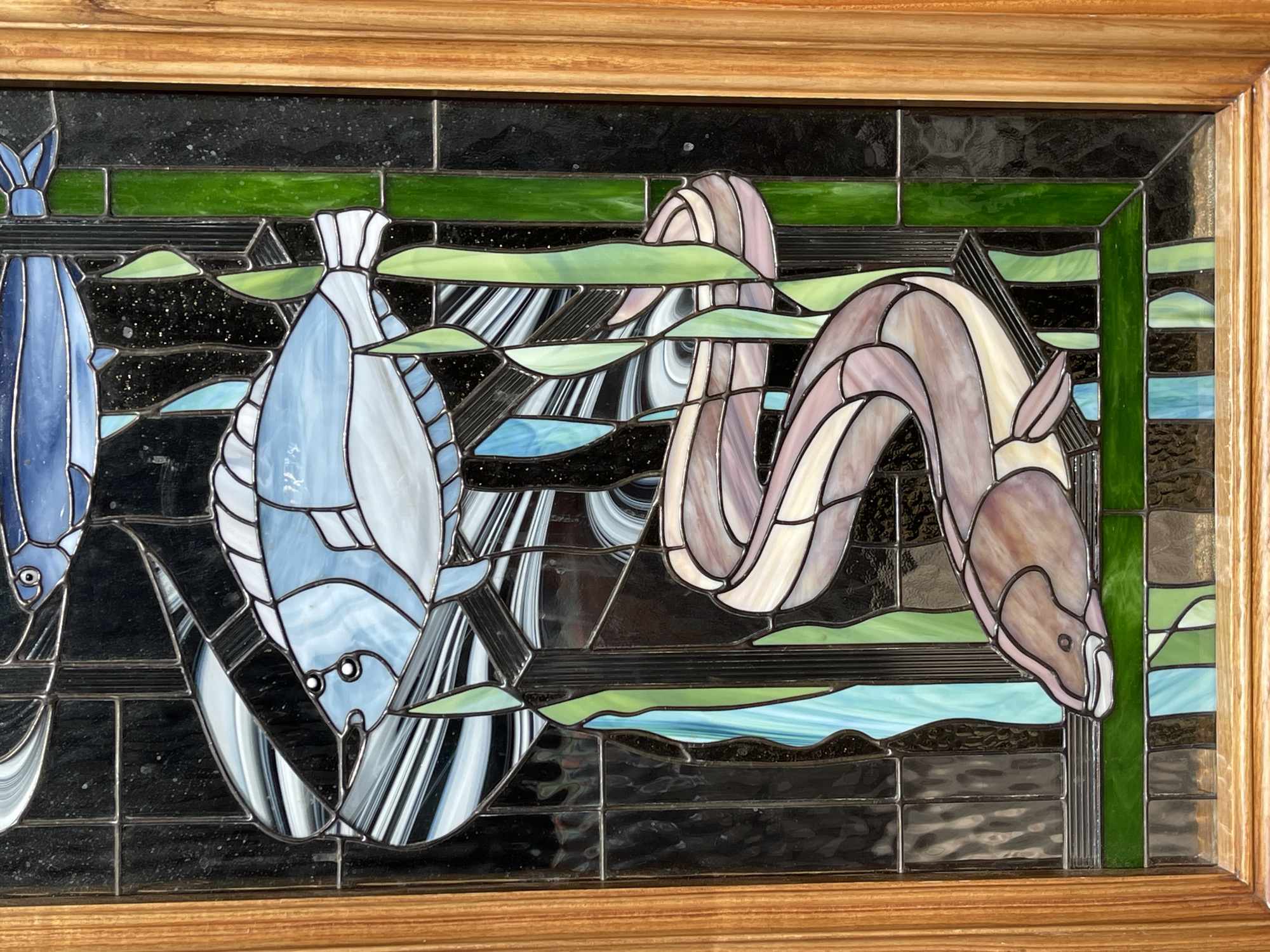 This is a stained glass panel depicting an underwater scene. The primary subjects are two fish and an eel. The fish, rendered in shades of blue and white, are positioned on the left side of the panel. One fish is larger and more detailed, with visible fins, eyes, and a patterned body. The other fish, partially visible, is a lighter blue. A long, segmented eel with a serpentine body in purple and white hues curves through the center and right side of the panel.
The background is composed of black, textured glass, simulating the dark depths of the ocean. Interspersed within this darkness are wavy bands of green and light blue glass, representing aquatic plants or water currents. Streaks of black and white glass create a sense of movement and depth, possibly depicting water or refracted light.
The stained glass panel is framed by a wooden border, suggesting it is an art piece meant for display, possibly as a window or decorative panel. There is no visible text in the image. The scene is static, with no discernible activity beyond the representation of the aquatic life. The overall impression is one of stylized underwater beauty, with a focus on the forms and colors of the marine creatures. No specific time of day, weather, or emotional cues are evident.
