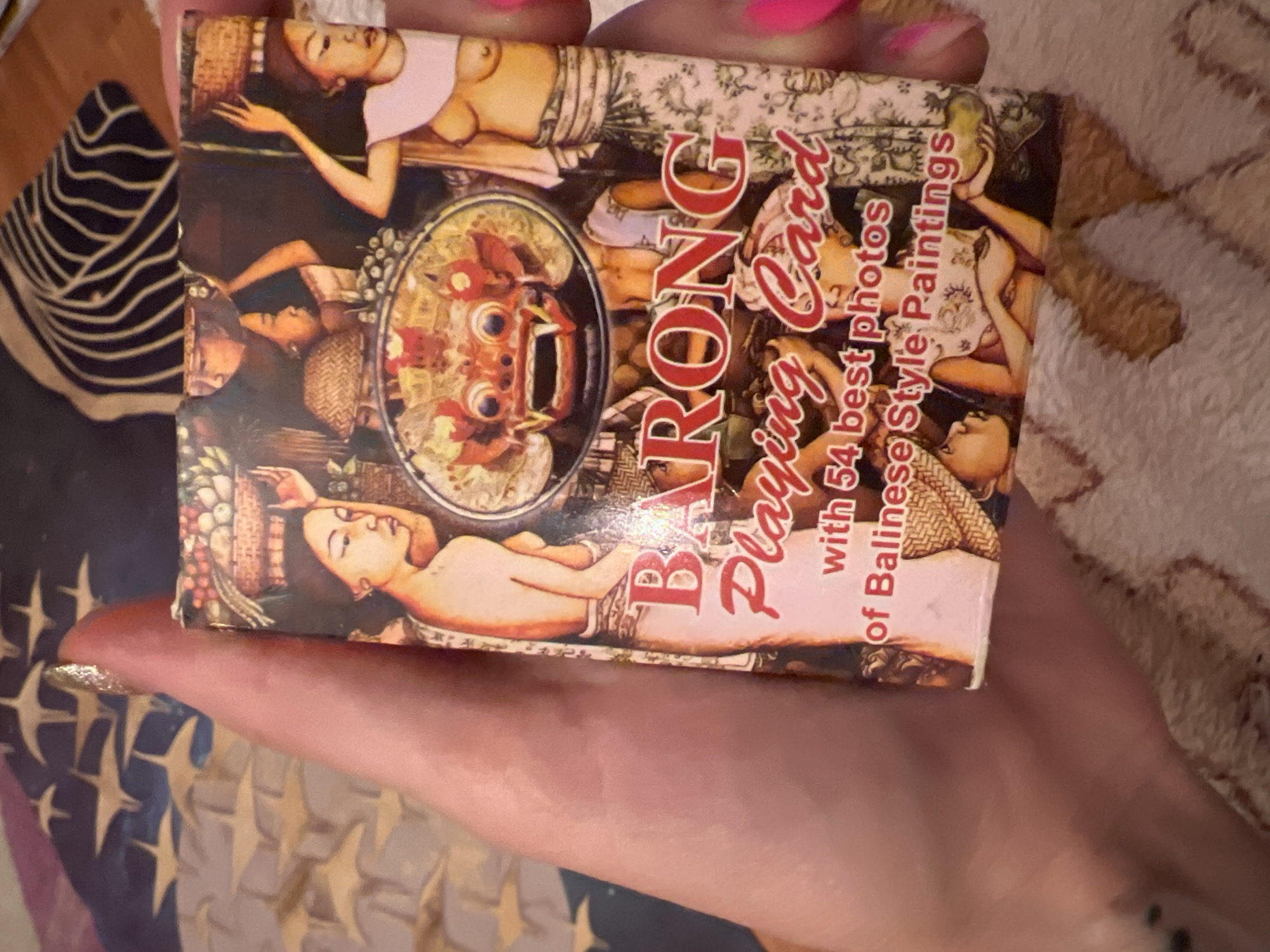 A hand holds a deck of "Barong Playing Card" with images of Balinese style paintings. The deck features a central image of a Barong mask, surrounded by depictions of women carrying baskets and wearing traditional attire. The text on the deck indicates it contains "54 best photos of Balinese Style Paintings." The background of the image shows a textured surface, possibly a blanket or rug. The location is not identifiable within the city of Krasnoe-na-Volge, Russia.