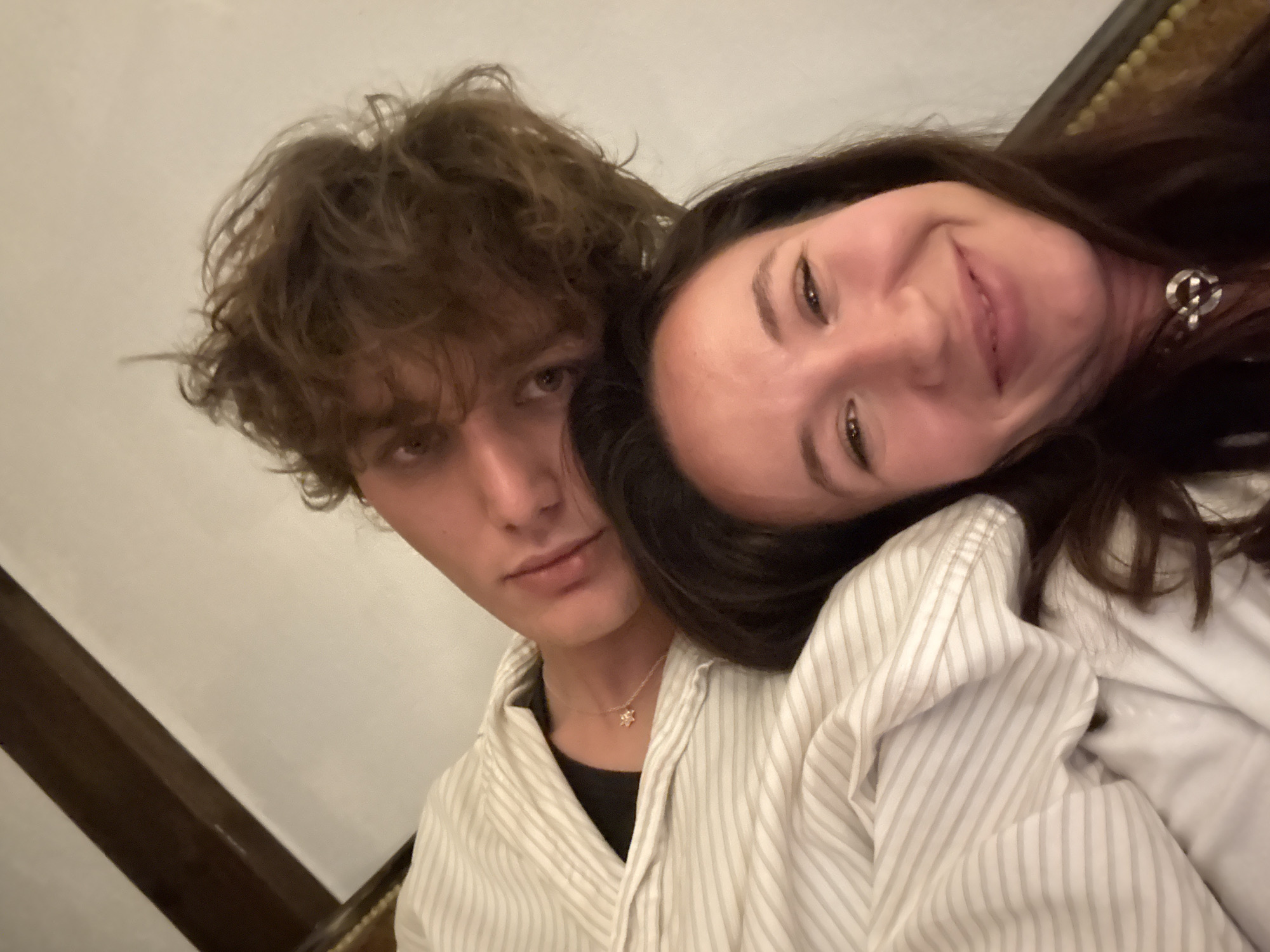A close-up shot depicts a young man and woman in an embrace. The man is on the left with tousled brown hair, fair skin, and a striped shirt over a black top. He wears a necklace. The woman is on the right, leaning her head towards the man. She has long dark hair, fair skin, and is also wearing a striped shirt. The lighting appears soft. The background is blurry and nondescript, showing a wall and wooden trim. There is no indication in the image that the location is Tel-Aviv, Israel.